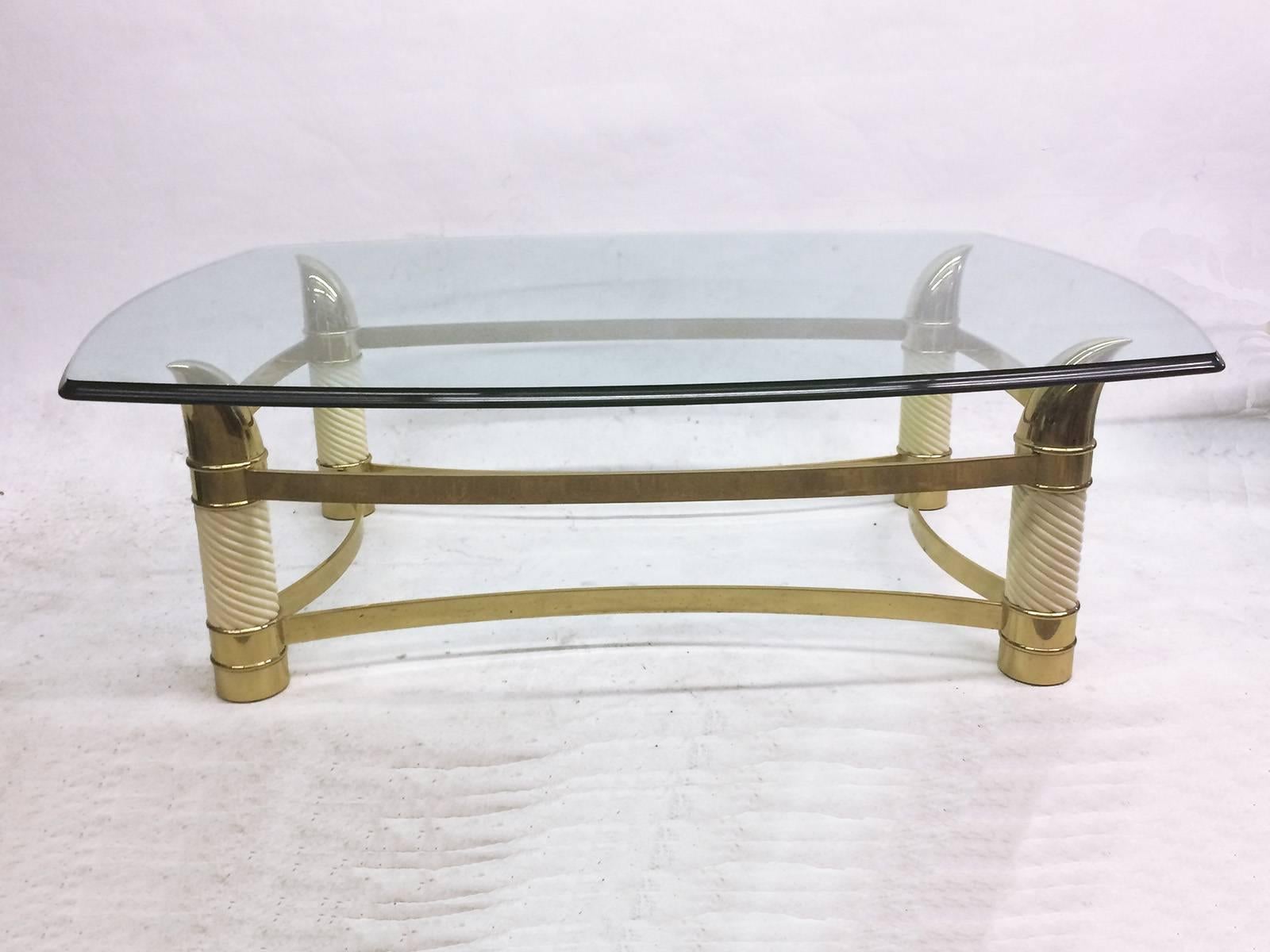 This Hollywood Regency coffee table is finished in white lacquer and brass. The beveled glass top surface rests on four brass horn-like supports that cantilever over the peaks. Four twisted columns on each corner have brass detailing at the base and