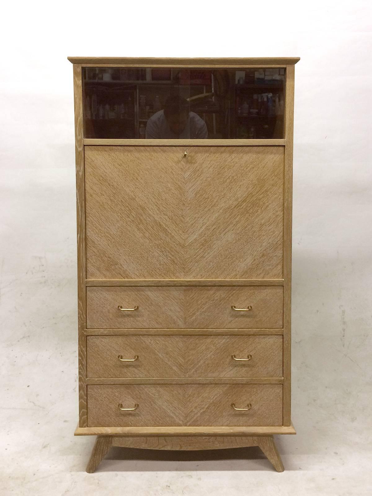 This Mid-Century secretaire is finished with bookmatched oak, brass fittings, and a light ceruse finish. The top section has sliding glass doors, the middle opens and has two drawers and one adjustable shelf and the bottom section has three drawers
