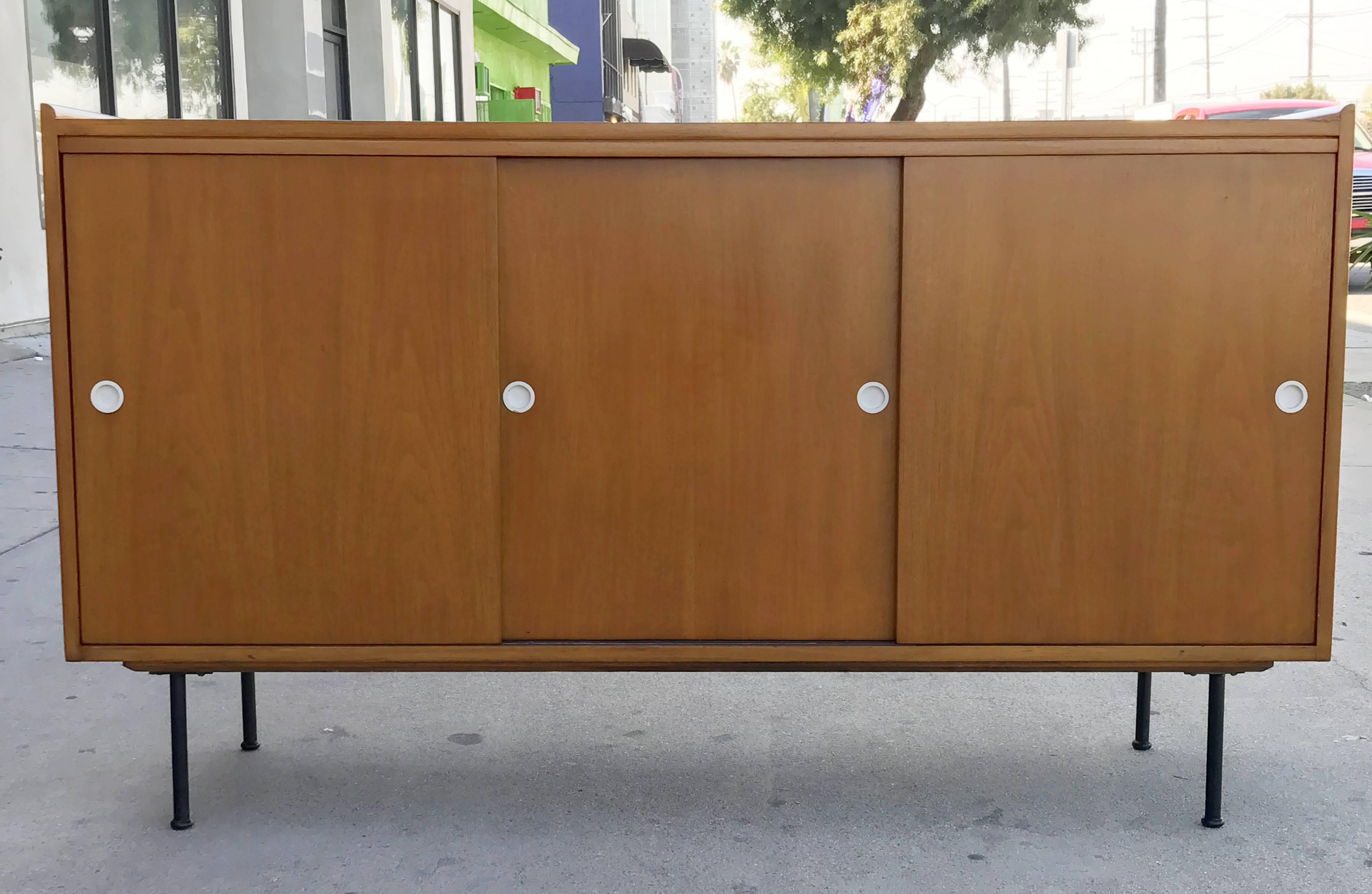 A sliding door credenza in the manner of American designer Paul McCobb. Walnut wood with blackened steel legs and white hardware. One section of the piece holds three drawers, the other a shelf.