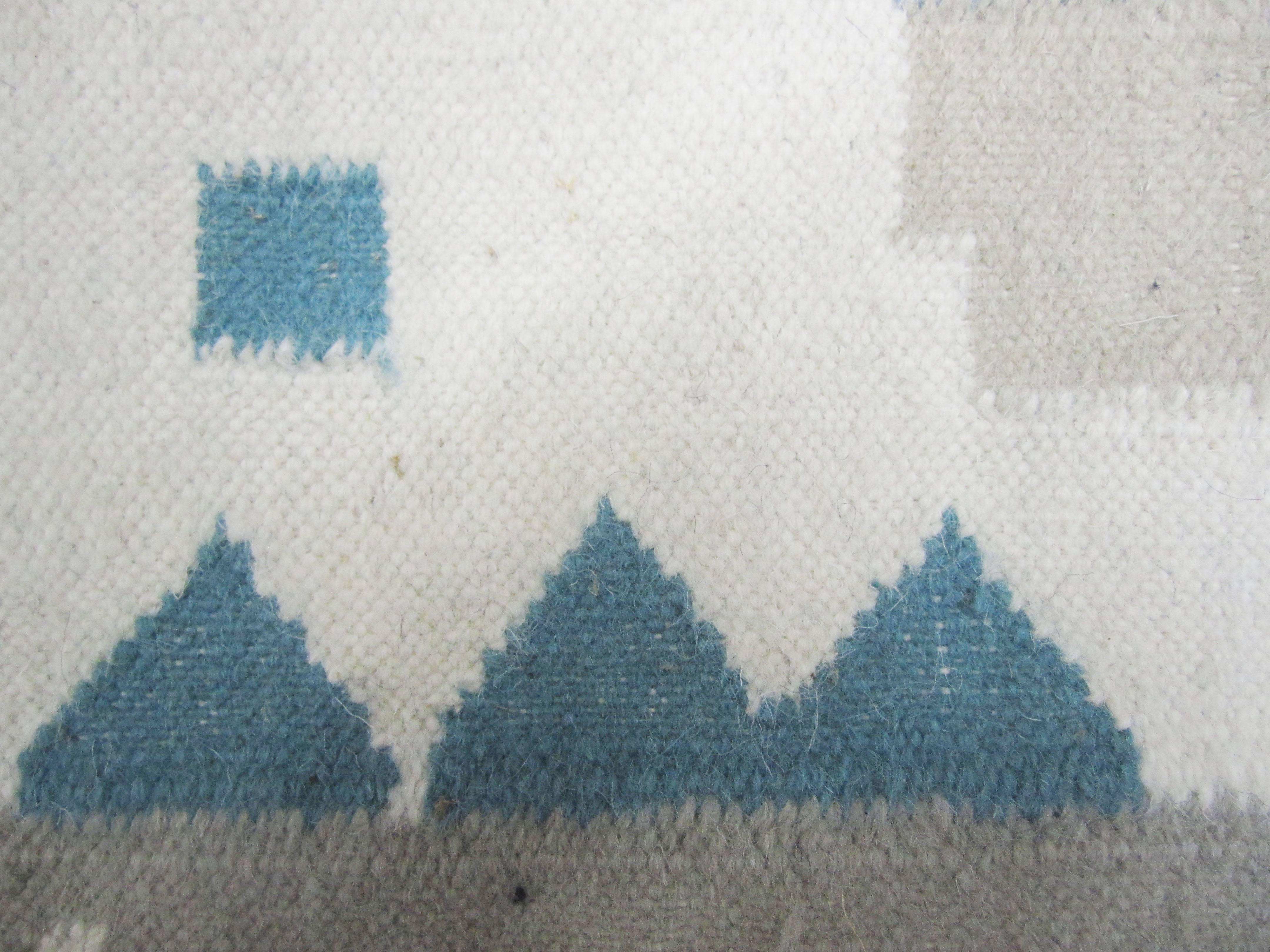 Woven wool rug or tapestry has a braided fringe on either side. Navy blue border with various blues and pastel shades. Likely depicts a Mexican landscape scene of a home with garden and mountains or ocean in the background.
 