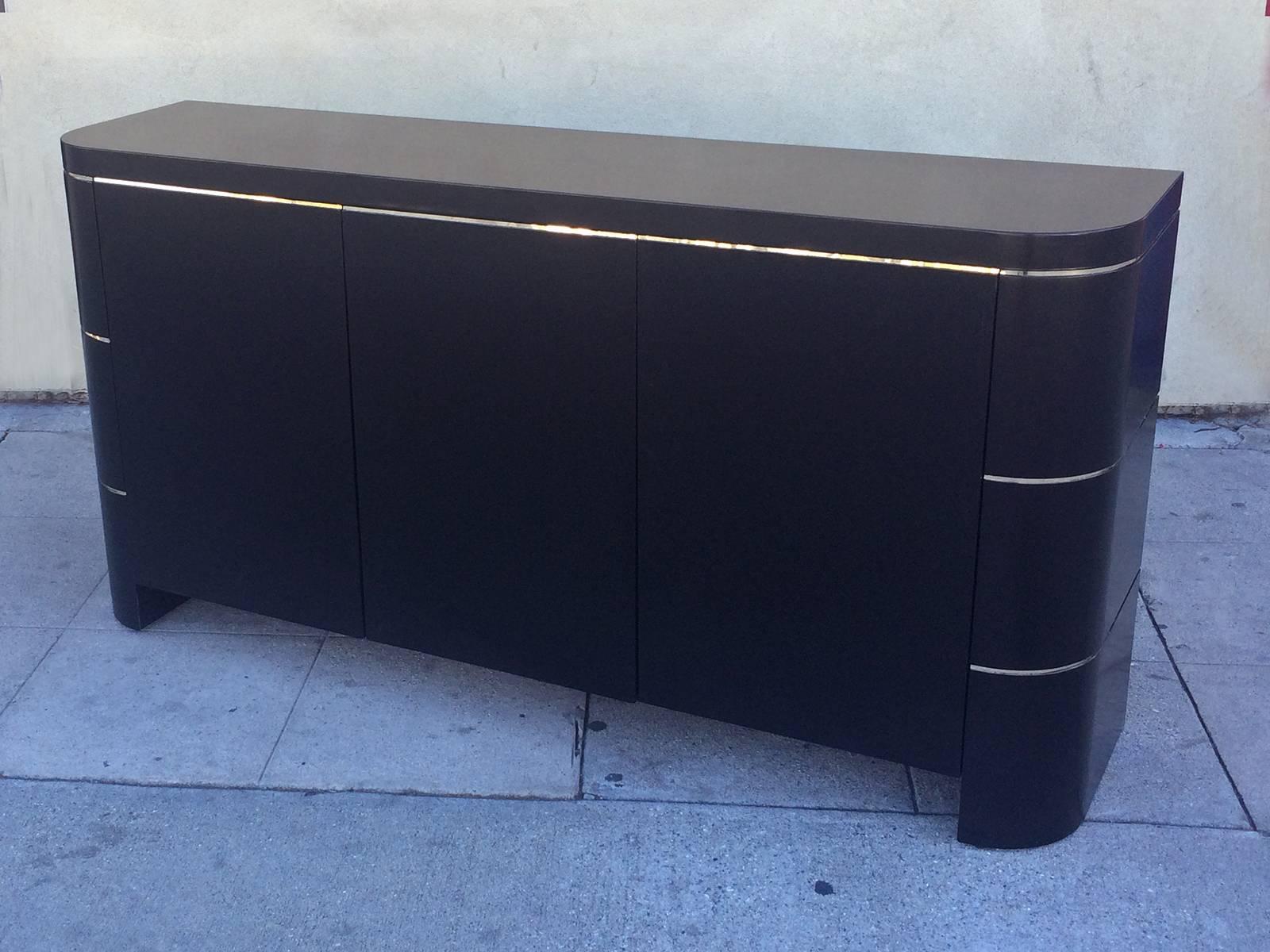 An elegant buffet featuring a finish in black lacquer with brass banding below top and in two other lower bands on ends. The rounded frame of this 1970s piece has three push-lock doors which open to reveal both shelf space and two pull-out drawers.