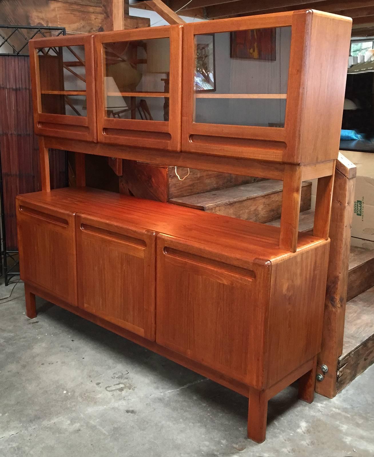 This a stacking Danish modern credenza and vitrine in teak. The piece has very modern lines and is finished with soft round edges. Both the vitrine and credenza are designed with built-in pulls. The vitrine features three individually adjustable