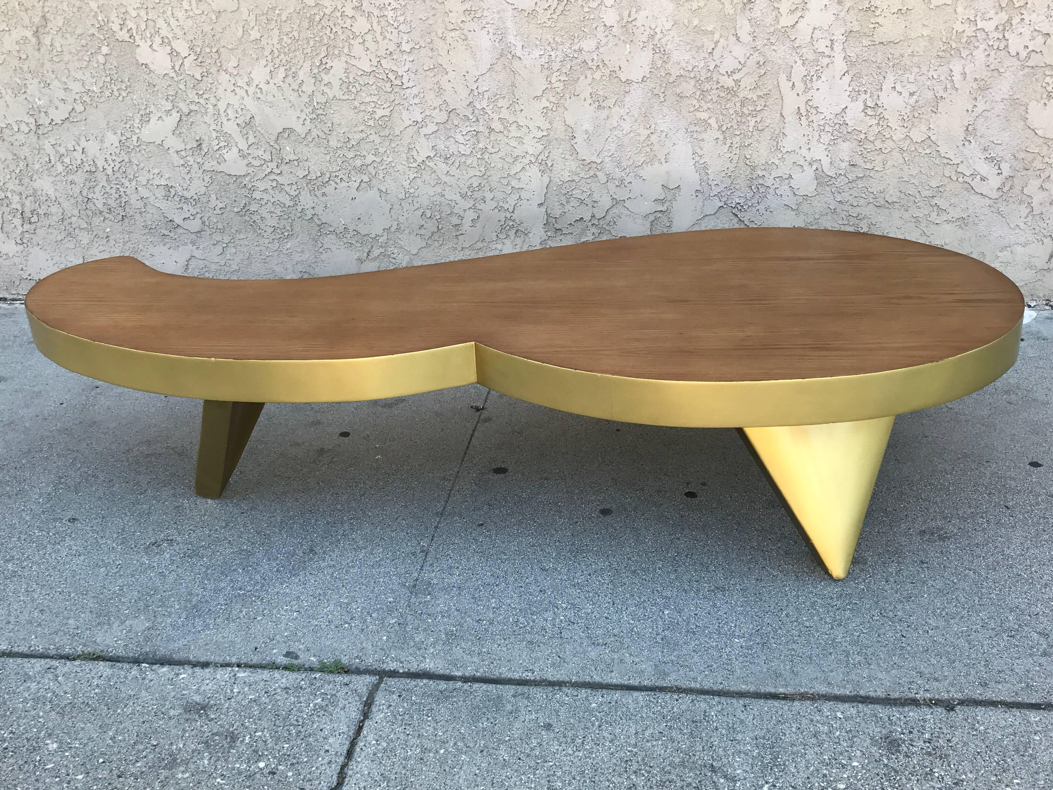 This coffee table features an interesting and contradictory mix of material: Oak with a flat finish for the top and gold lacquer for the skirt and the legs.
The shape is unusual and typically 1980s. (The fun and whimsical part).

The wood top is