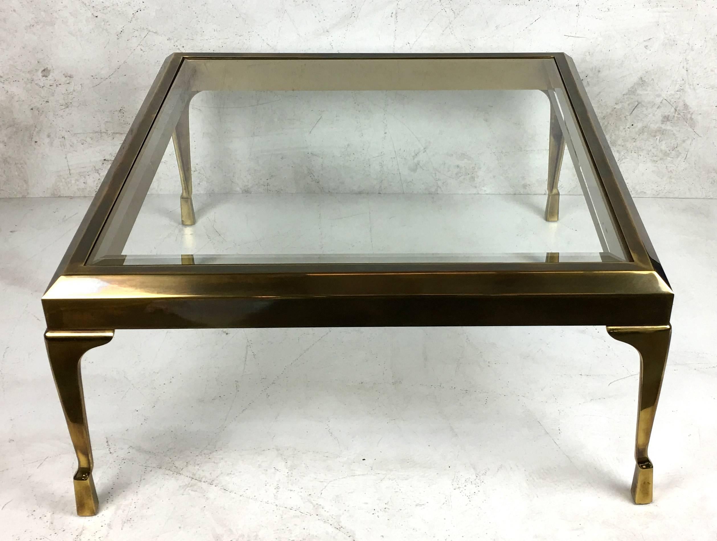 Sophisticated brass coffee table raised on sculpted brass legs with stylized hooved feet by Mastercraft. The brass has its beautiful original antiqued finish along with its bevelled glass tabletop intact and in excellent condition. These designs by