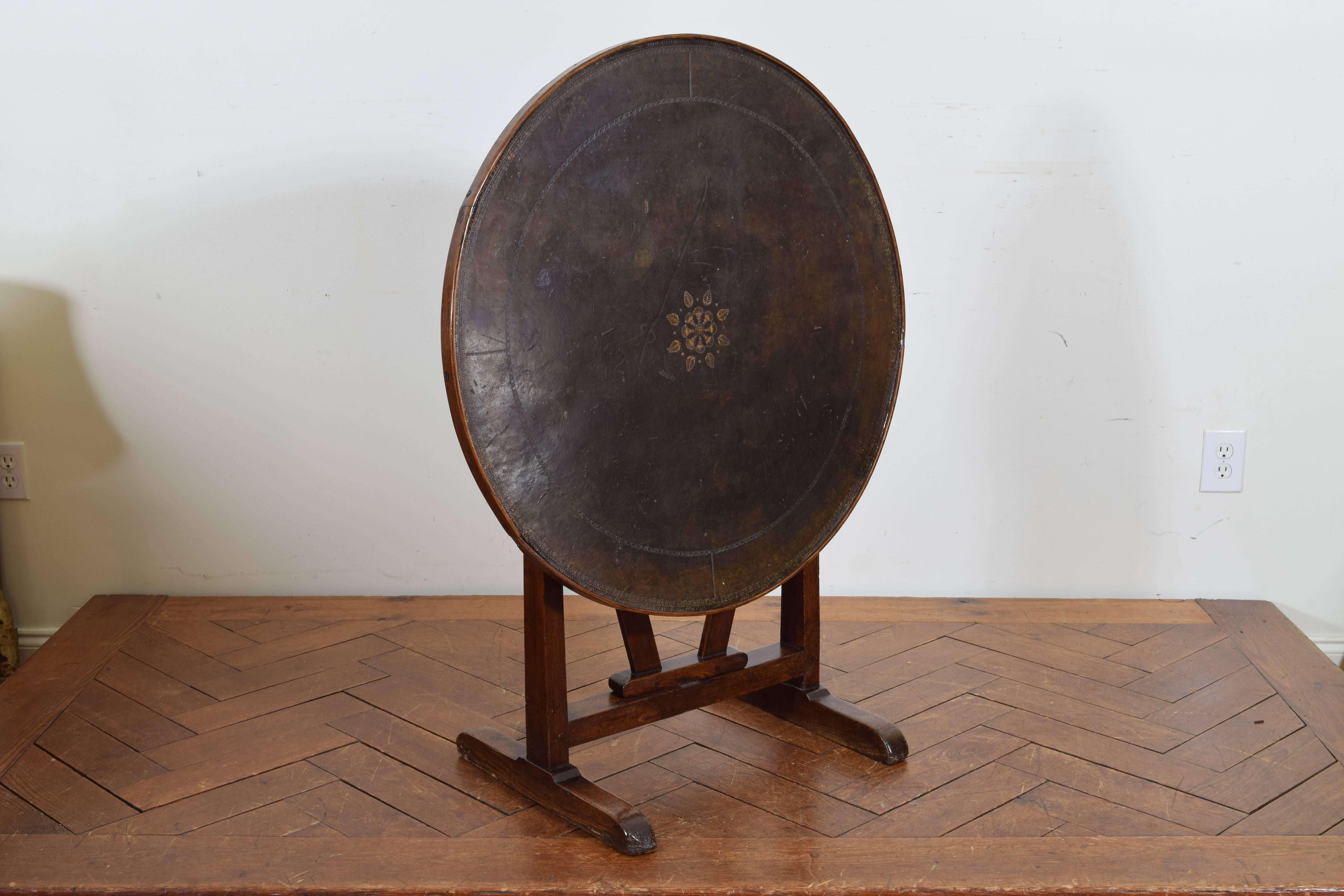 The circular top covered in tooled and painted leather, folding with a centre rotating support and trestle-form legs with bracket feet.