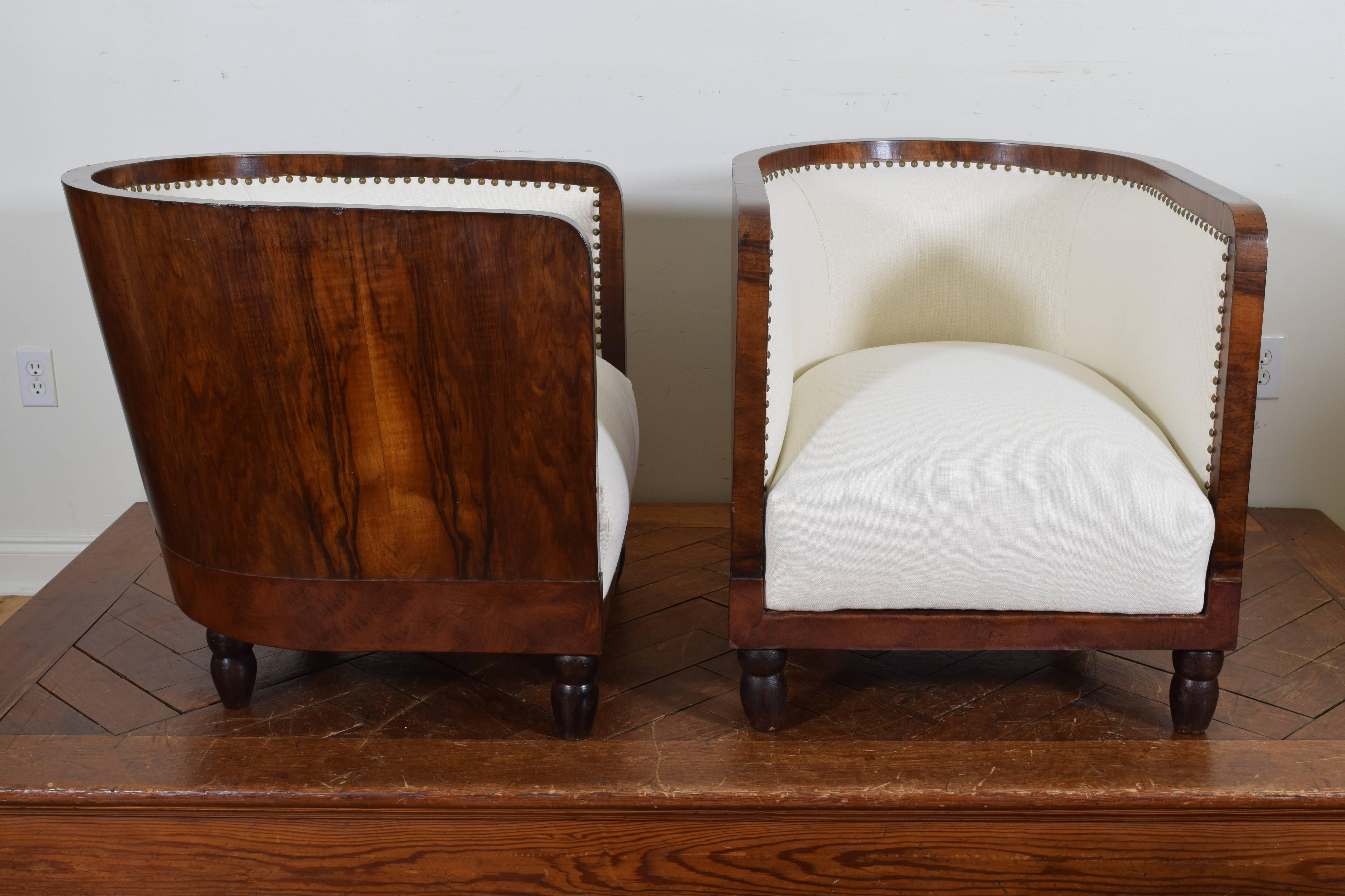 Having deep seats upholstered in off-white linen and covered entirely in walnut veneer, the lower section of each with a leather band, raised on turned ebonized feet.