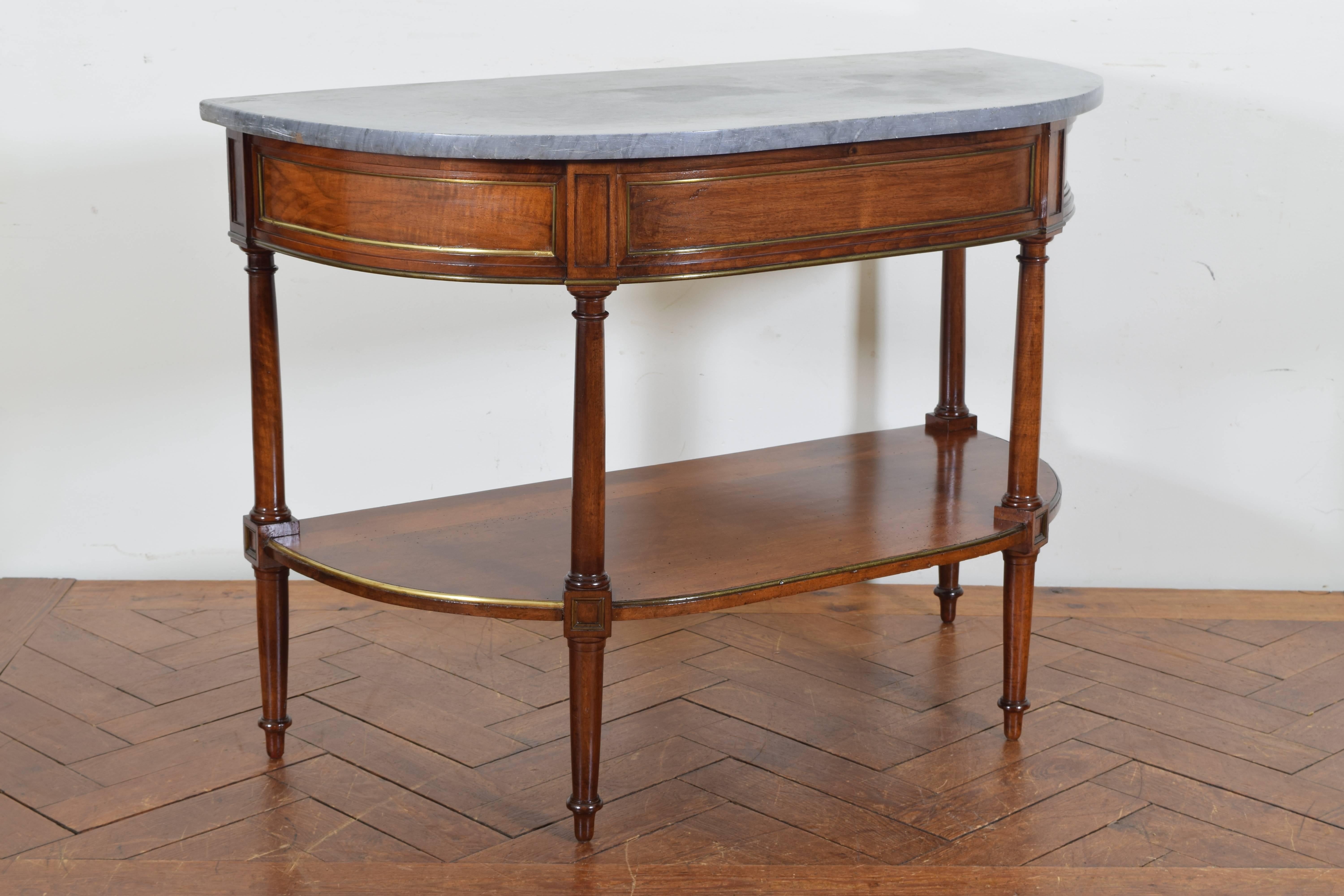The grey marble-top of demilune form above a conforming frame, the panels of the upper section trimmed in brass, having turned legs and tapered feet, the lower section also trimmed in brass.