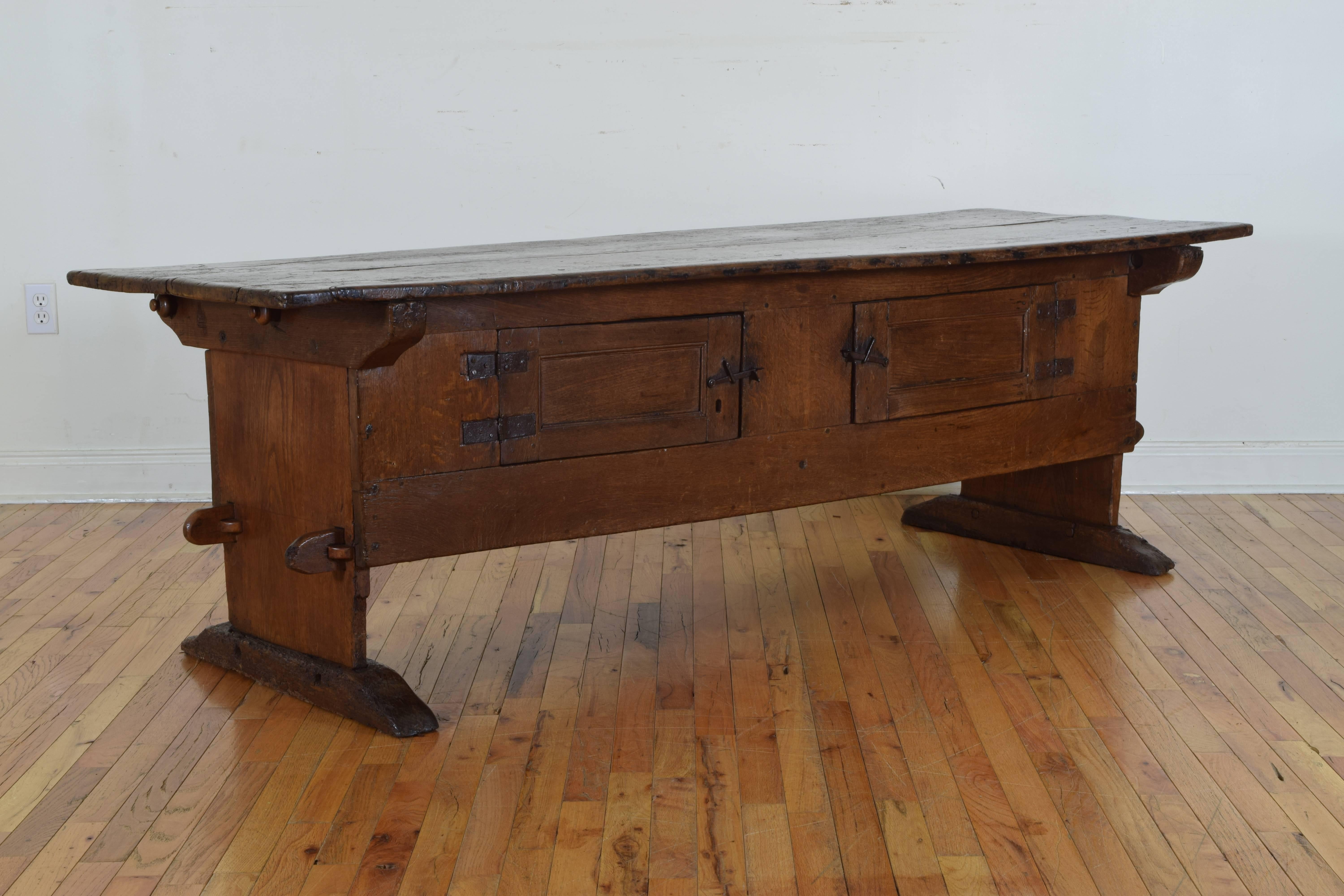Constructed entirely of a light oak, the table has a two board rectangular top above a conforming cabinet housing two hinged doors, the cabinet supported by trestle legs on elongated bracket feet, probably originally used in a kitchen for an all