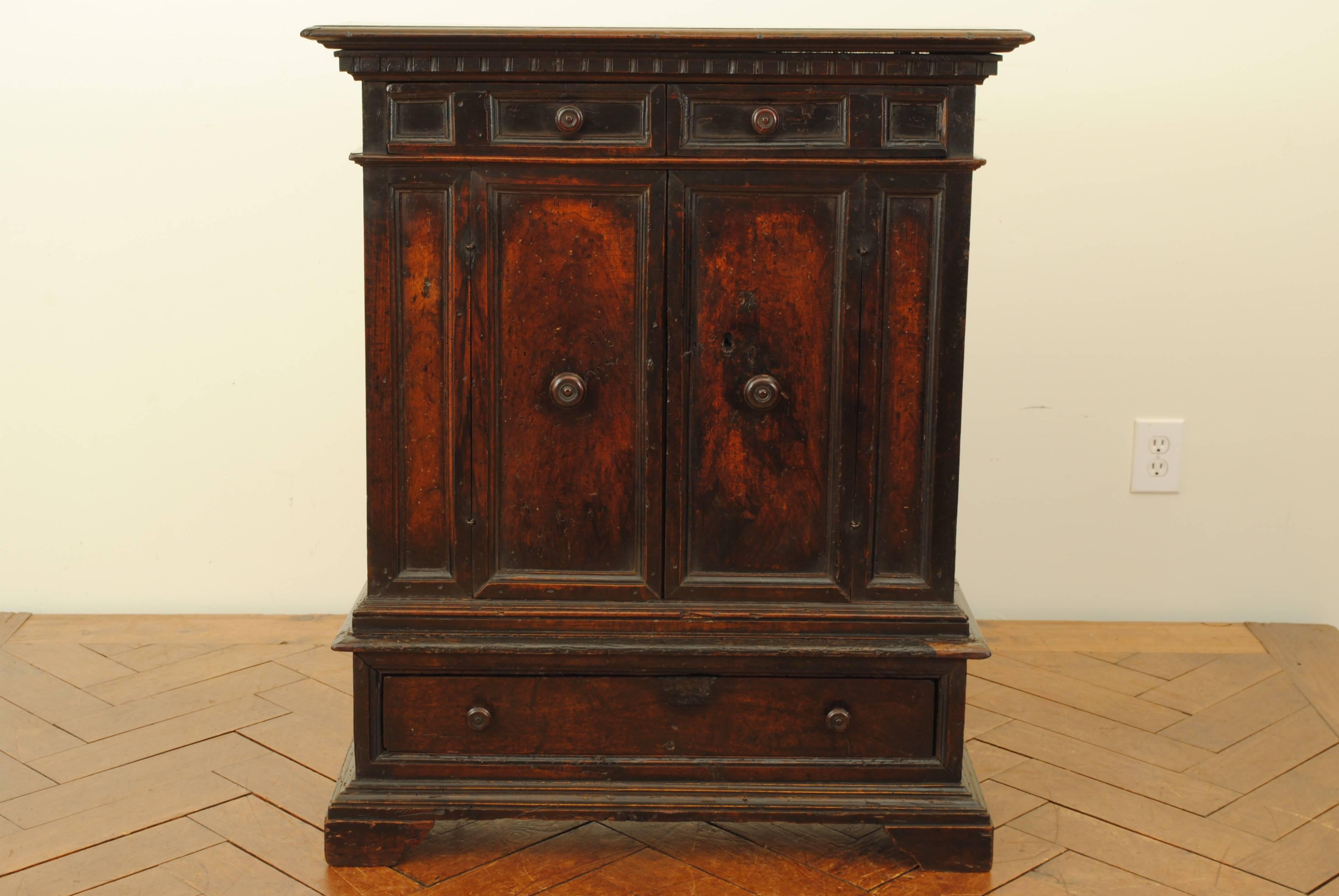 With a rich and dark waxed patina, having a rectangular top with molded edge and a dentile carved lower edge, the conforming case housing two upper drawers above two doors and a single larger bottom drawer, paneled throughout and raised on a plinth