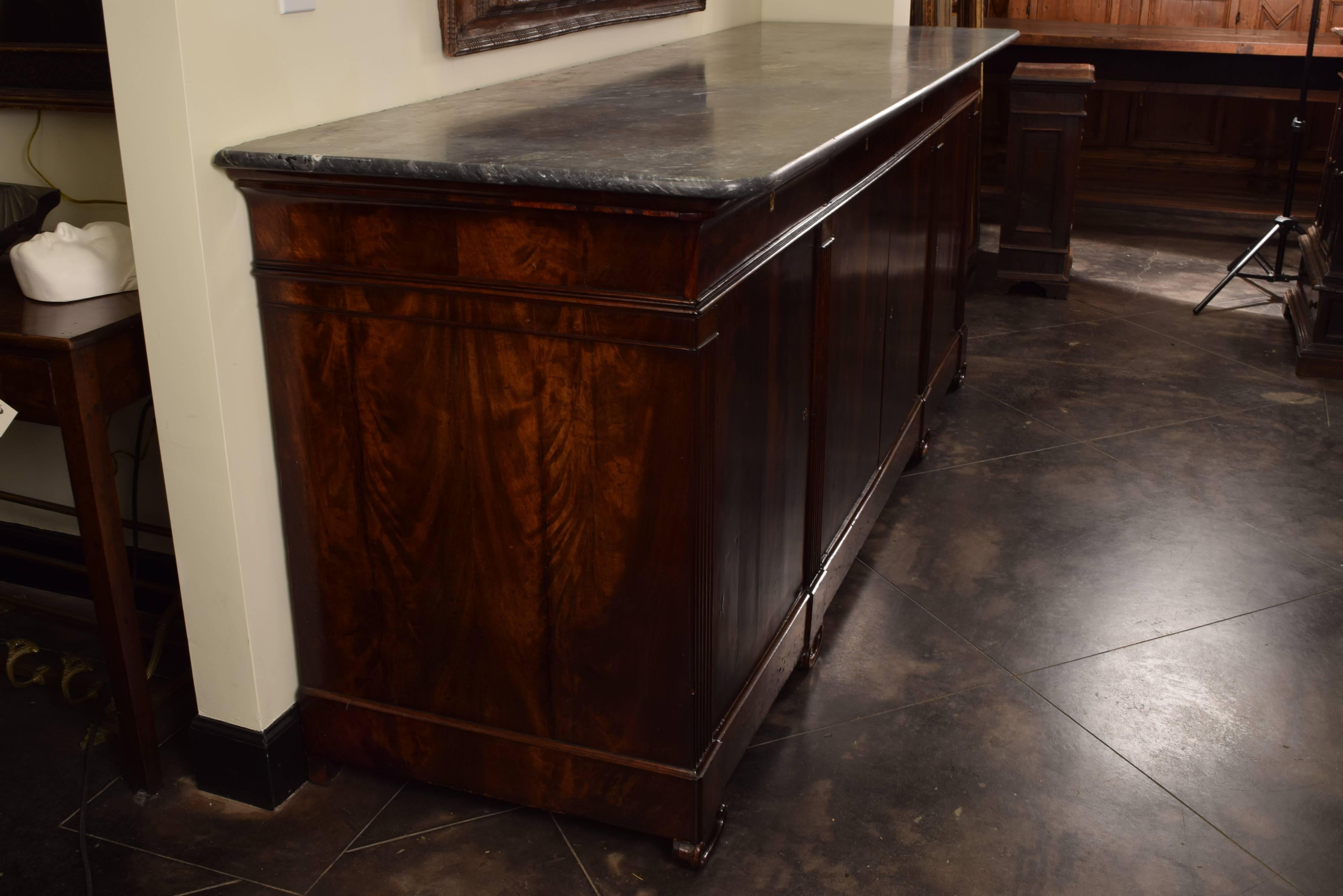 The rectangular marble-top with rounded front corners above a conforming case veneered in bookmatched mahogany, housing four drawers over four doors, the doors separated by stylized fluted columns, raised on scroll carved feet, all doors and drawers