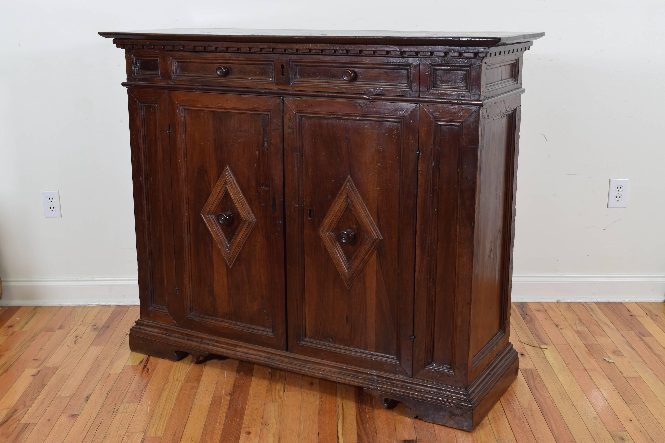 Credenza of shallow depth, the rectangular top with a dentil molded apron above a conforming case with paneled sections housing one drawer over two doors with raised lozenge panels and wooden pulls, raised on a graduated plinth base and bracket feet.