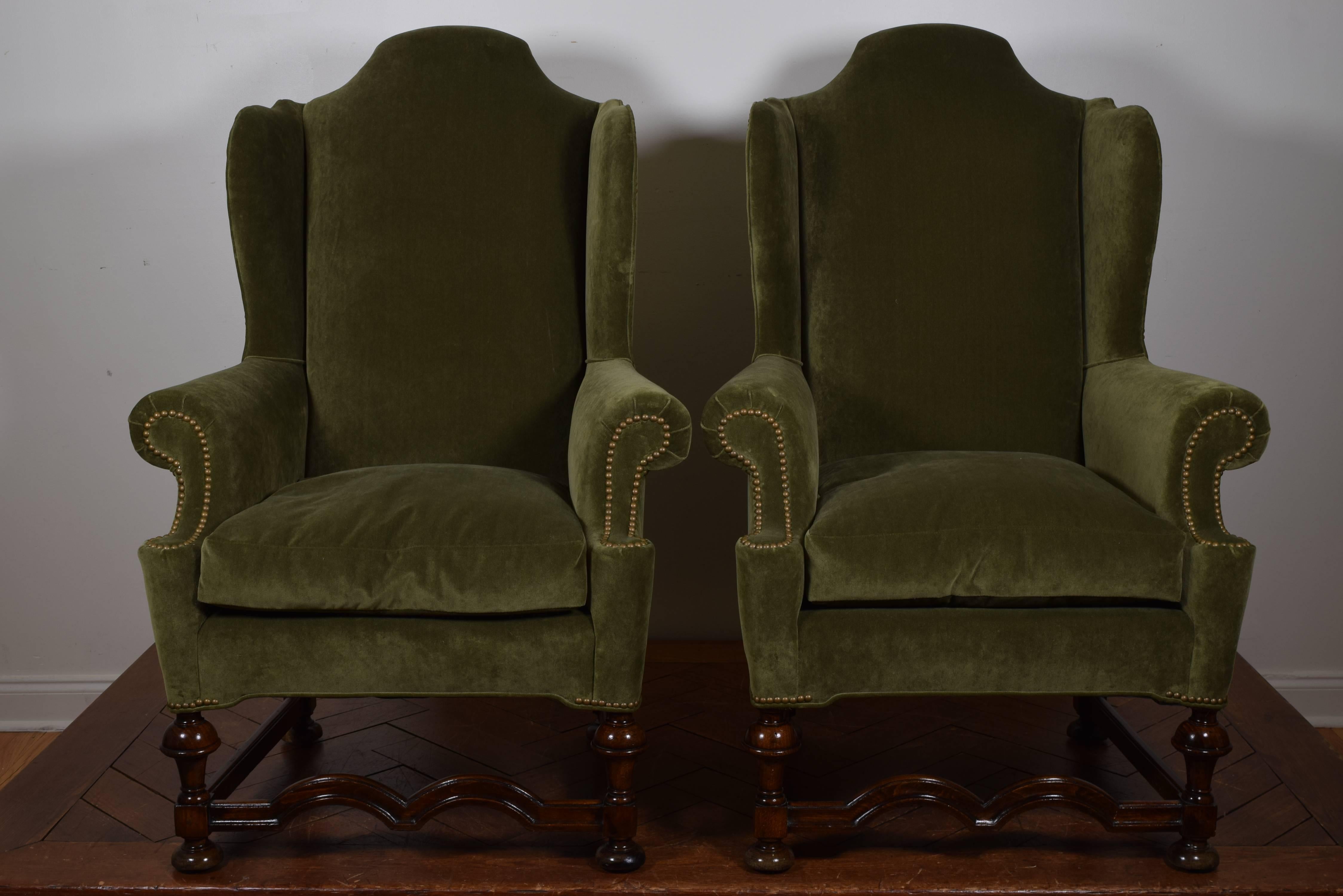 The arched backrests behind fully upholstered wings and rolled arms, the upholstery trimmed in brass nailheads, down seat cushions, the frames nicely carved with turned legs and channeled stretchers, raised on bun feet.