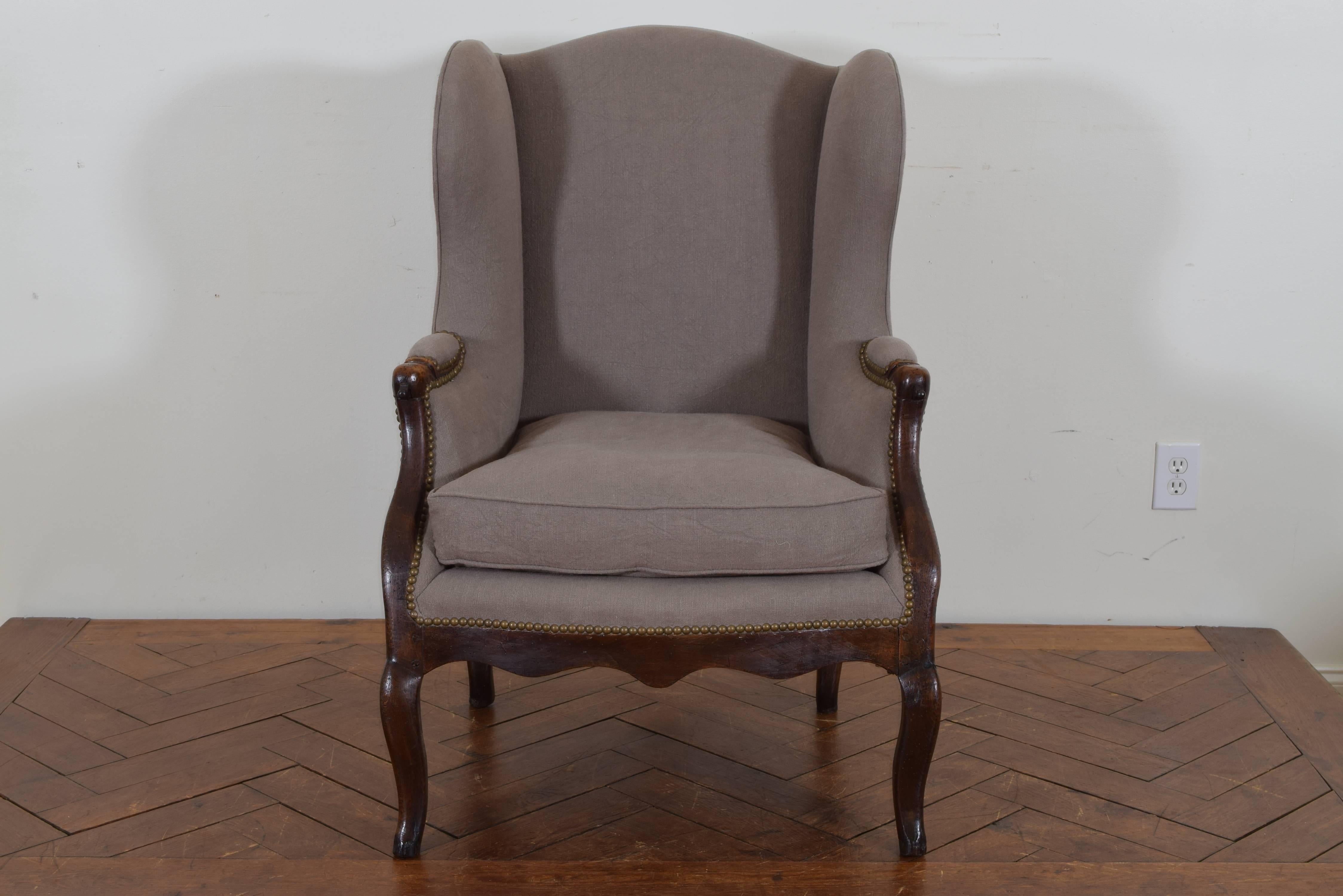 The arched backrest and shaped wings upholstered in a grey linen and trimmed in patinated brass nailheads, the shaped arms, legs and apron in walnut, with iron slides in the arms which would have supported a tray.