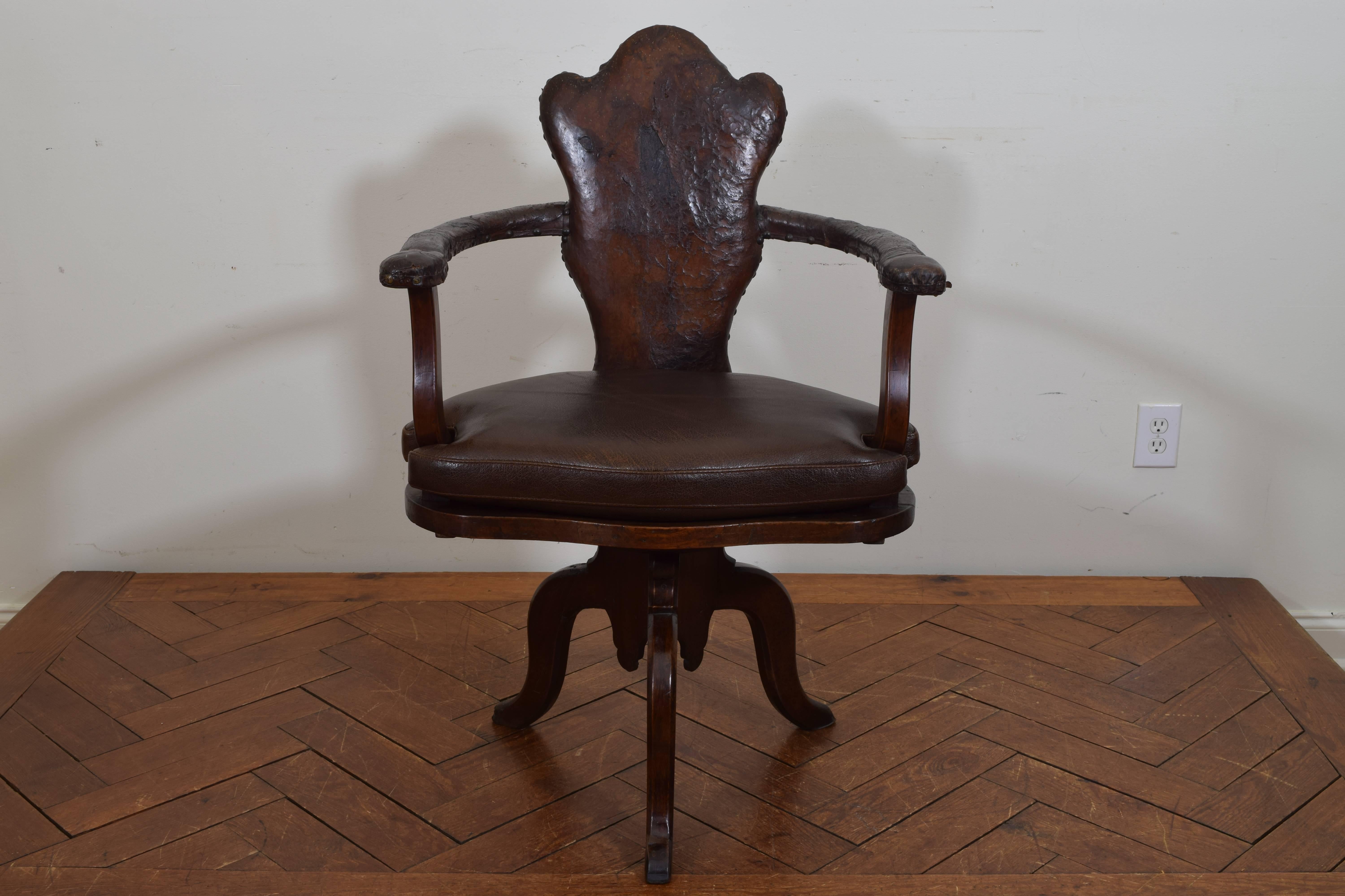 Having a shaped backrest and U-shaped arms raised on shaped supports, all upholstered in antique leather and trimmed in nailheads, the generous seat upholstered in a new leather hide and resting atop a round swiveling deck sitting above a carved