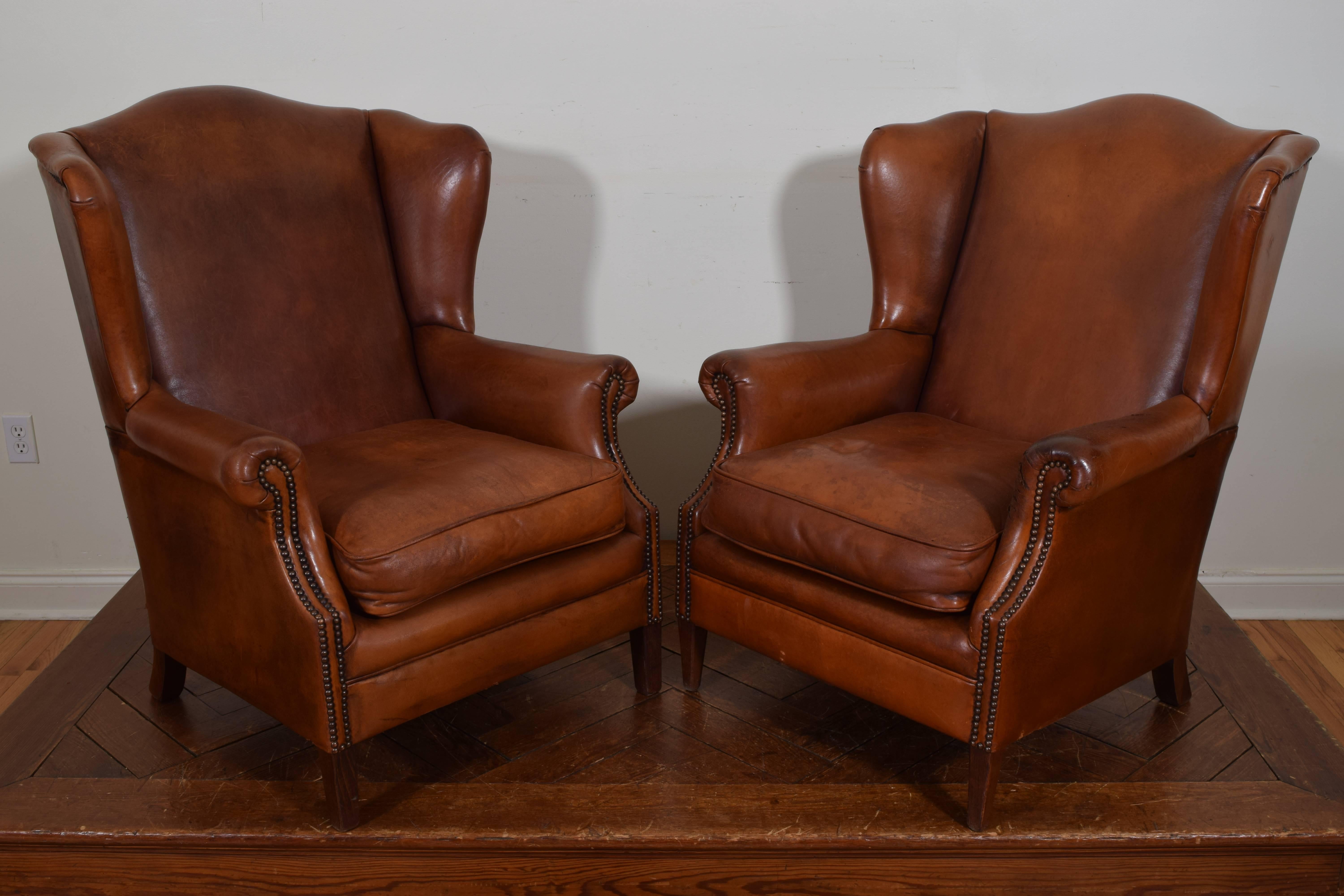 The pair having arched backrest and enclosed shaped wing sides, trimmed entirely in patinated brass nailheads, having loose cushions, raised on slightly different feet, one chair having front feet more tapered.