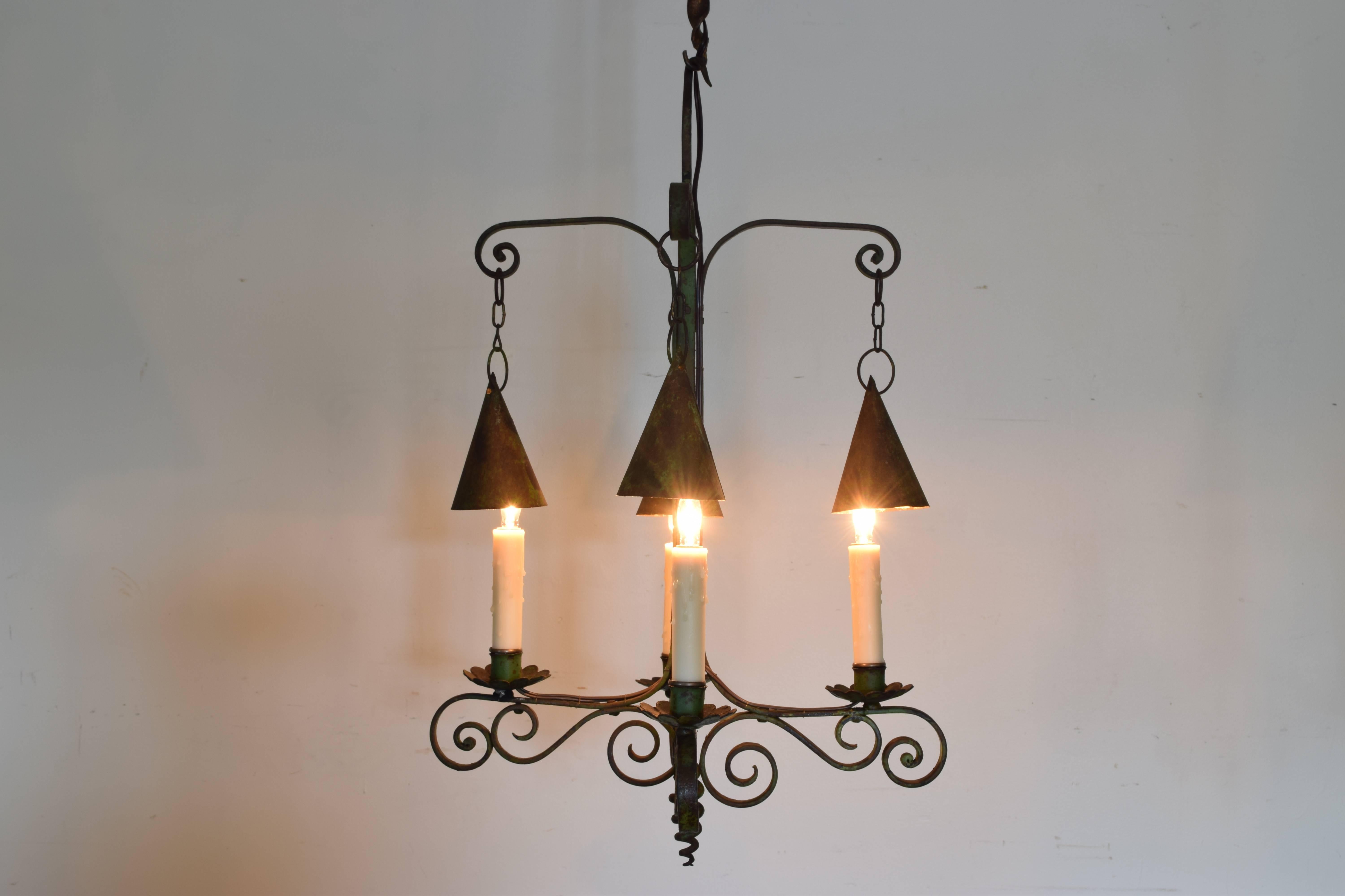 The fixture having four arms, each light with a hanging conic lantern shade above it, the body made up scrolled iron.