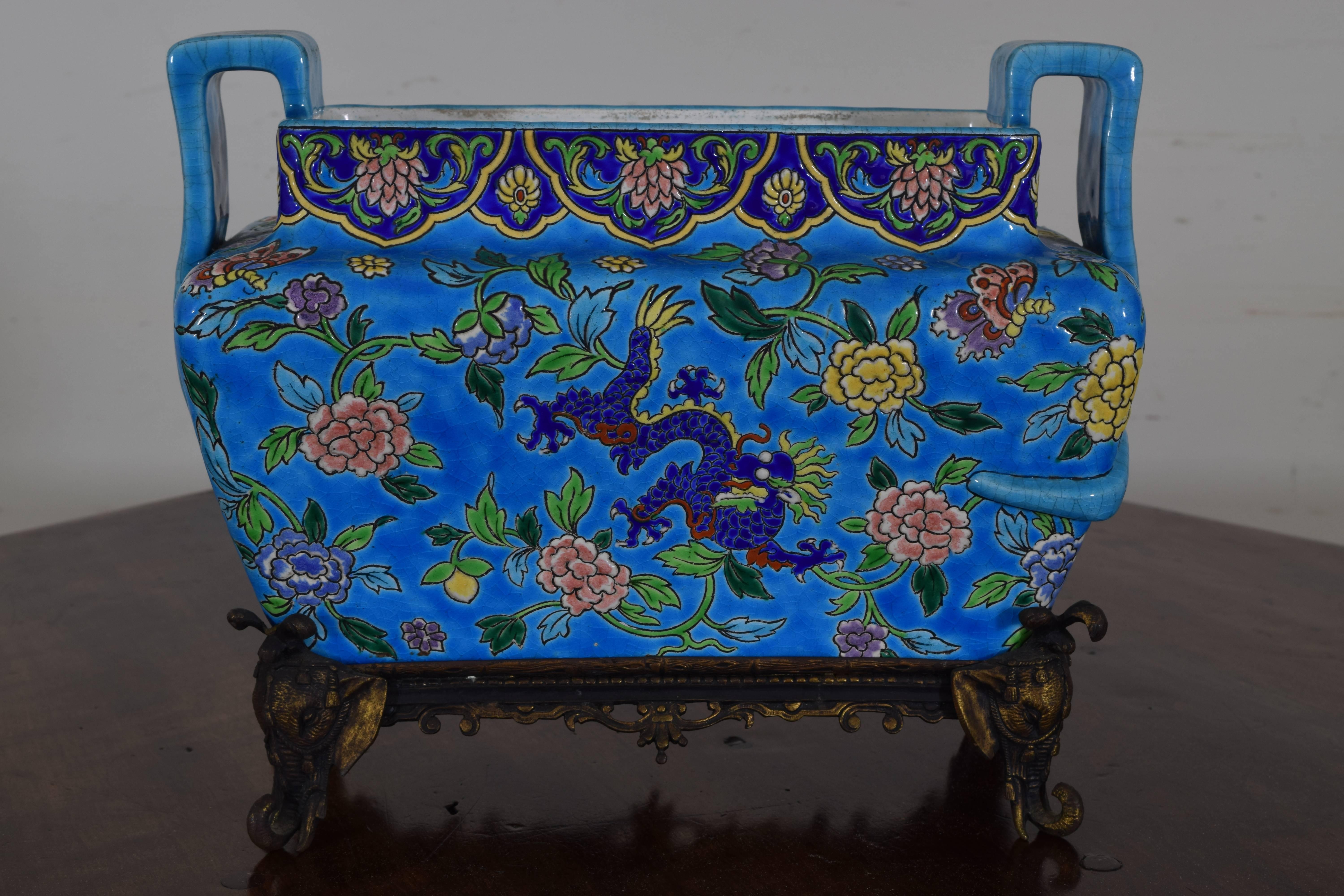 Of continental origin and having a makers mark on underside, glazed cachepot in brilliant colors of blue with dragons and various colored flowers, the gilt metal stands with elephants heads.