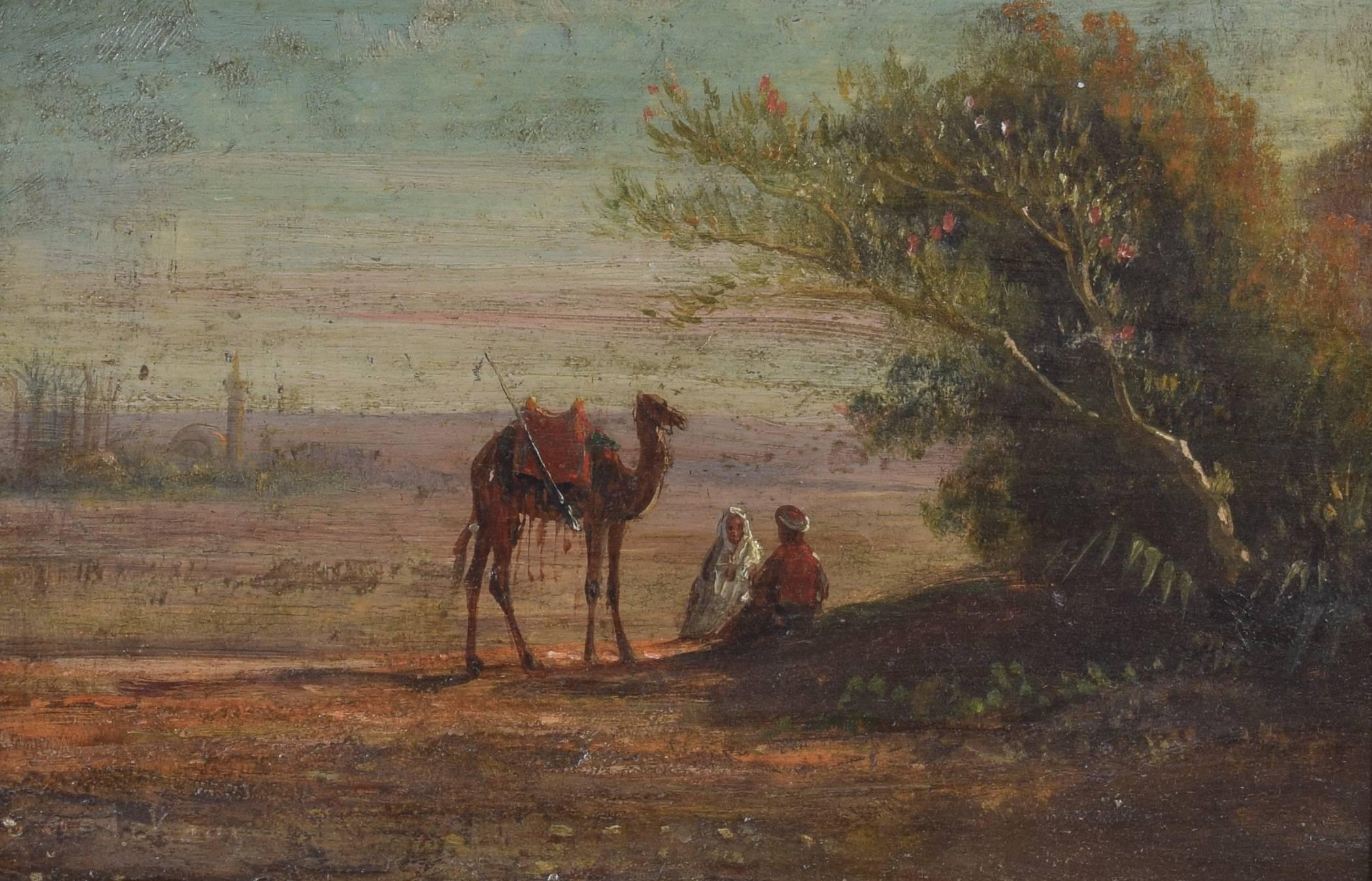 likely French and painted in the orientalist manner, the scene depicting two travelers and a camel at an oasis, a city in the left background, the frame of carved walnut, first quarter of the 20th century.