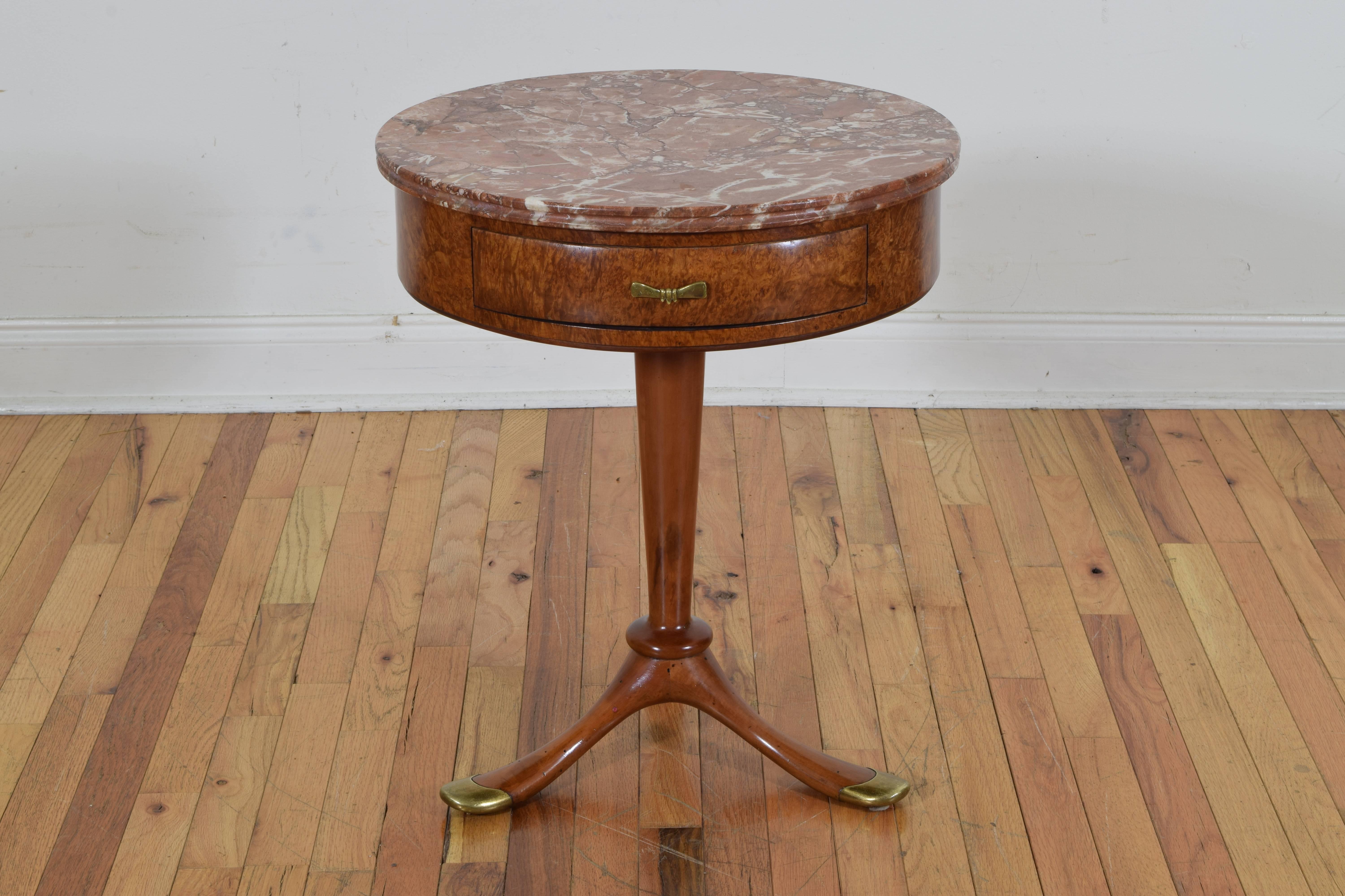 having a circular marble top with a channeled edge, the burl walnut veneered and solid walnut table having one drawer with a brass handle, raised on a tapered central column terminating in a tripartite base issuing legs with brass castors 