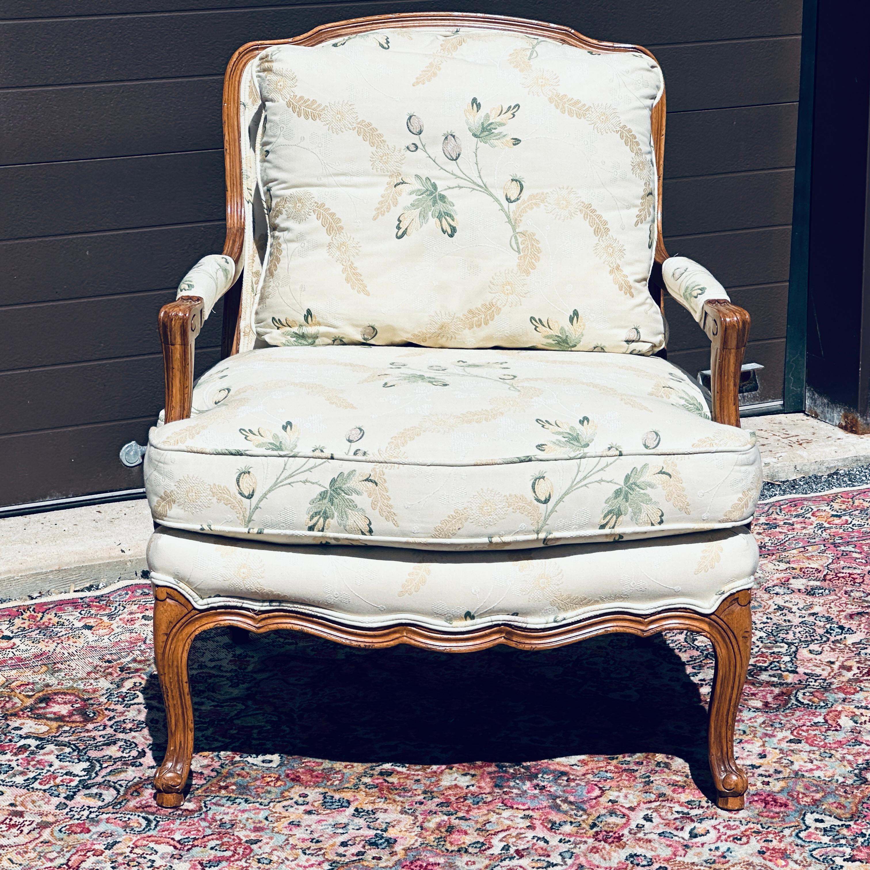 High end French style open arm bergere chair by Baker Furniture Company featuring a carved fruitwood frame with original floral upholstery. The loose seat and back cushions are flippable. The main color of the upholstery is Cream.