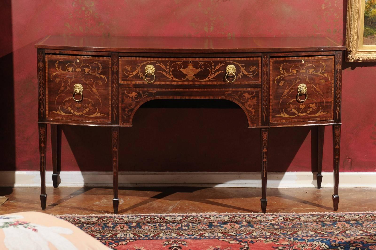Exquisite mahogany bowfront sideboard adorned with beautiful marquetry inlay on the front and down the tapered legs. Satinwood banding is on the top as well as the sides. There is a centre long drawer and two doors on either side. Lion head pulls.