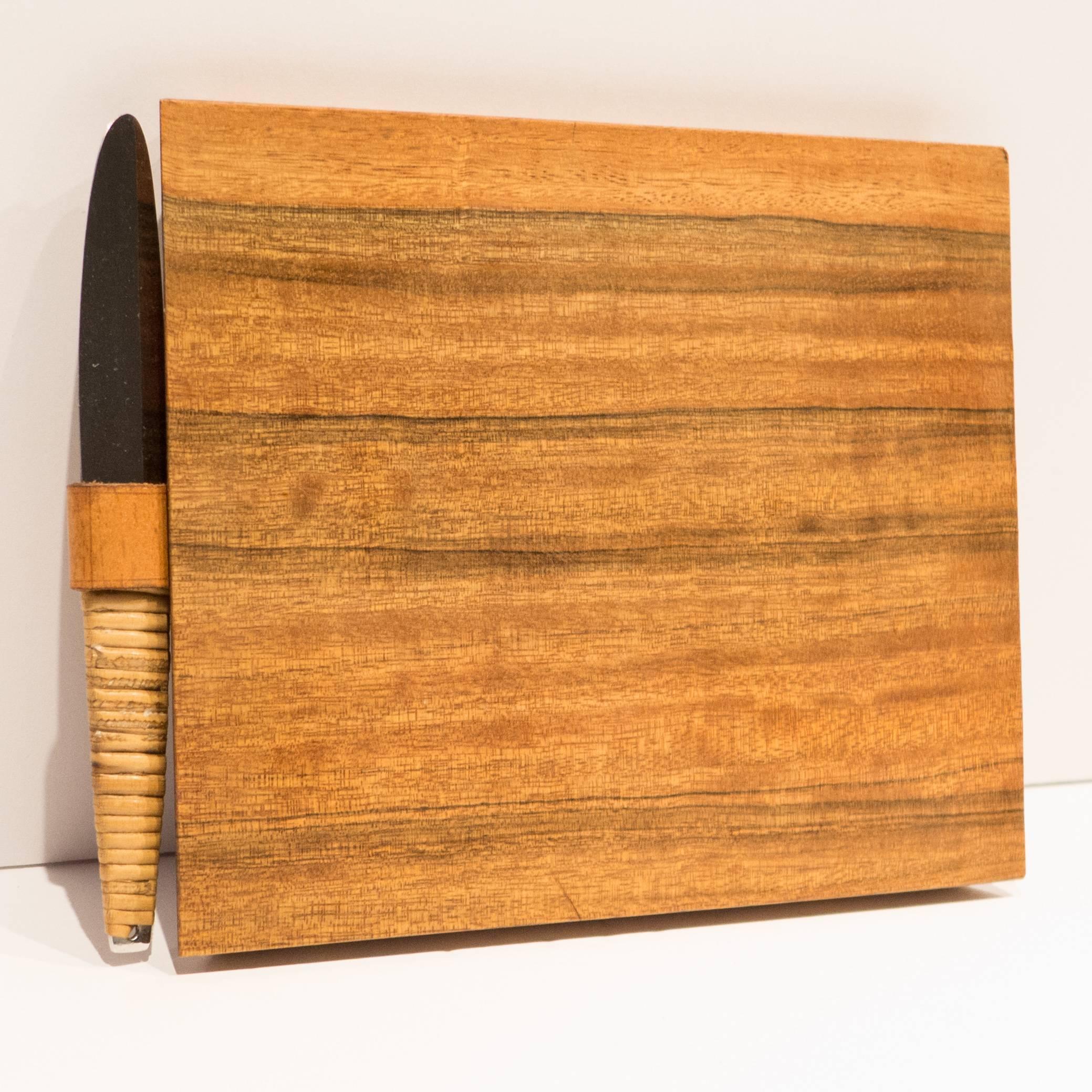 Wooden cutting board in a trapezoidal shape with a leather loop holding a cane-wrapped stainless steel knife. The knife is marked 