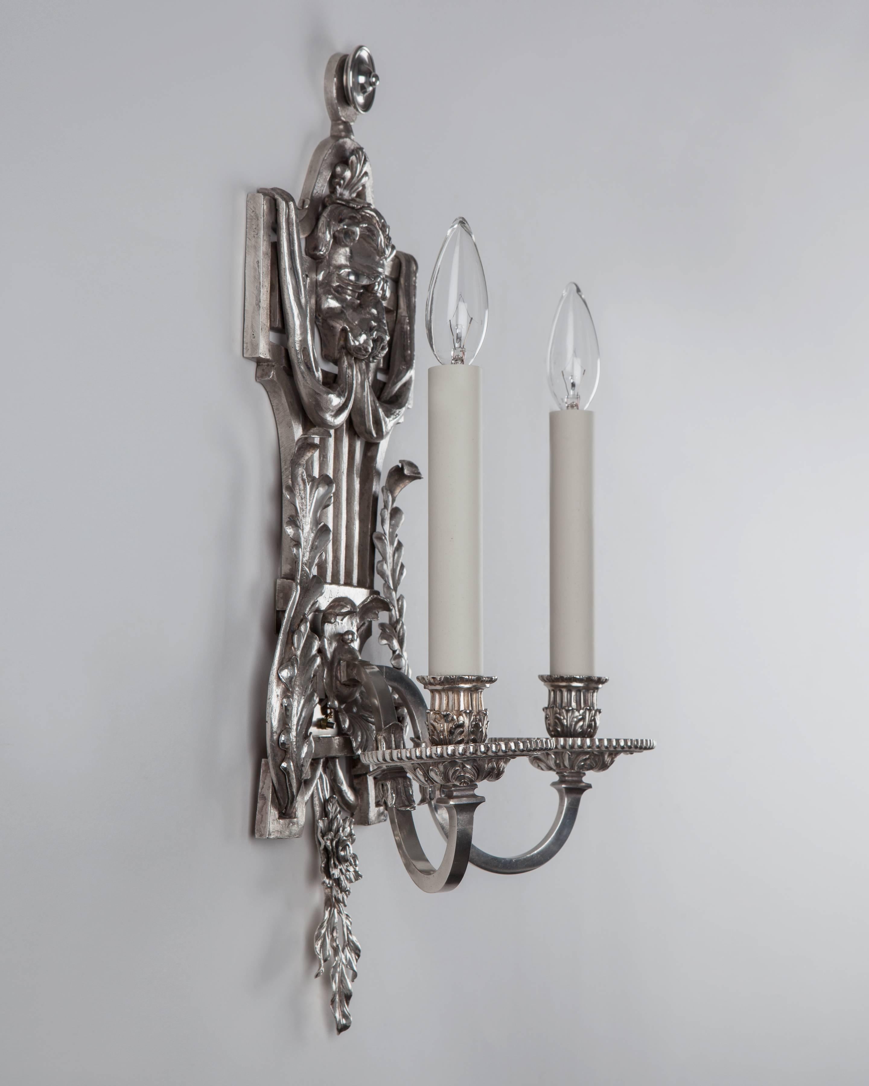AIS3019
A pair of two arm lyre-back sconces in a worn silver plate finish. Square-section arms spring from openwork back plates detailed with foliate patterns crowned with a lion's head relief. Attributed to the New York maker E. F. Caldwell Co.