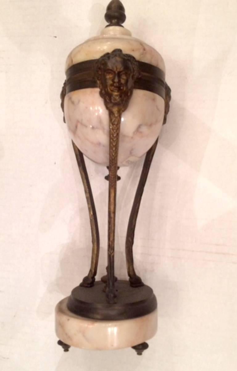 Urne aus Marmor und Bronze, mit Masken und einem dreibeinigen Sockel.
 
Maße: 12