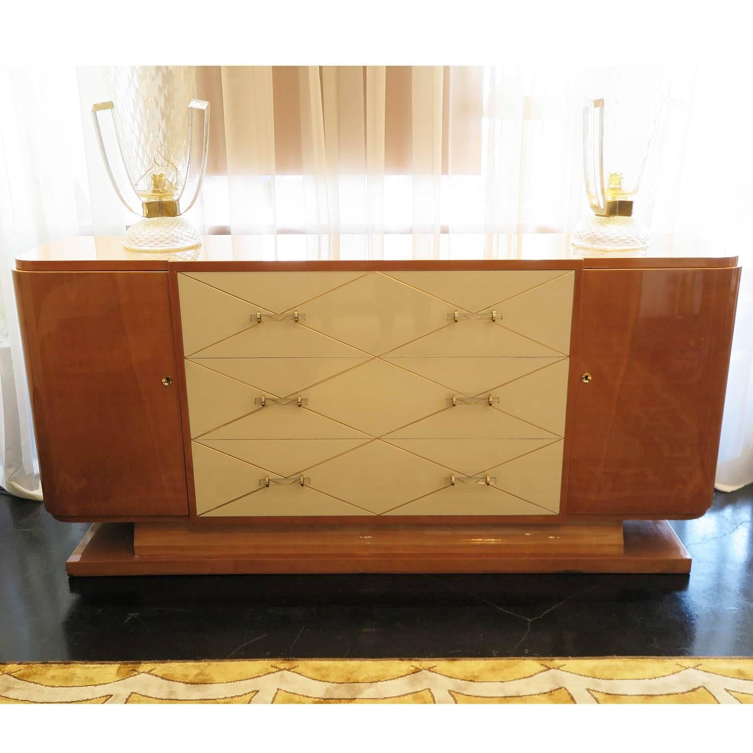 Glamorous warm-tone maple veneer sideboard with ivory lacquered drawers and original Lucite and brass pulls. A gold leaf harlequin pattern decorates the front of the three drawers. The sideboard also features two locking cabinet doors that conceal