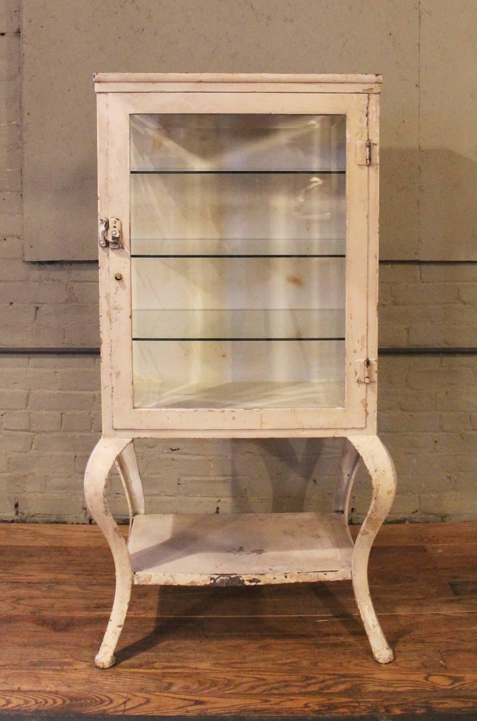 Vintage Industrial antique steel, metal and glass medical, pharmacy storage cabinet with three glass shelves and cabriole legs. Beautiful patina, worn ivory paint. Overall dimensions are 28" x 19 3/4" x 52 3/4". Bottom shelf measures