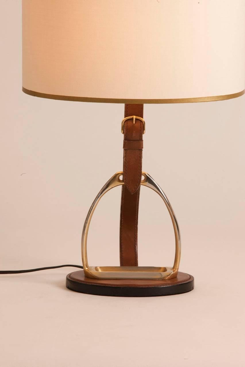 French Mid-Century leather and brass table lamp attributed to Longchamp. Lamp features equestrian detailing and a brass buckle and stirrup. The shade is custom and was made in Paris.
