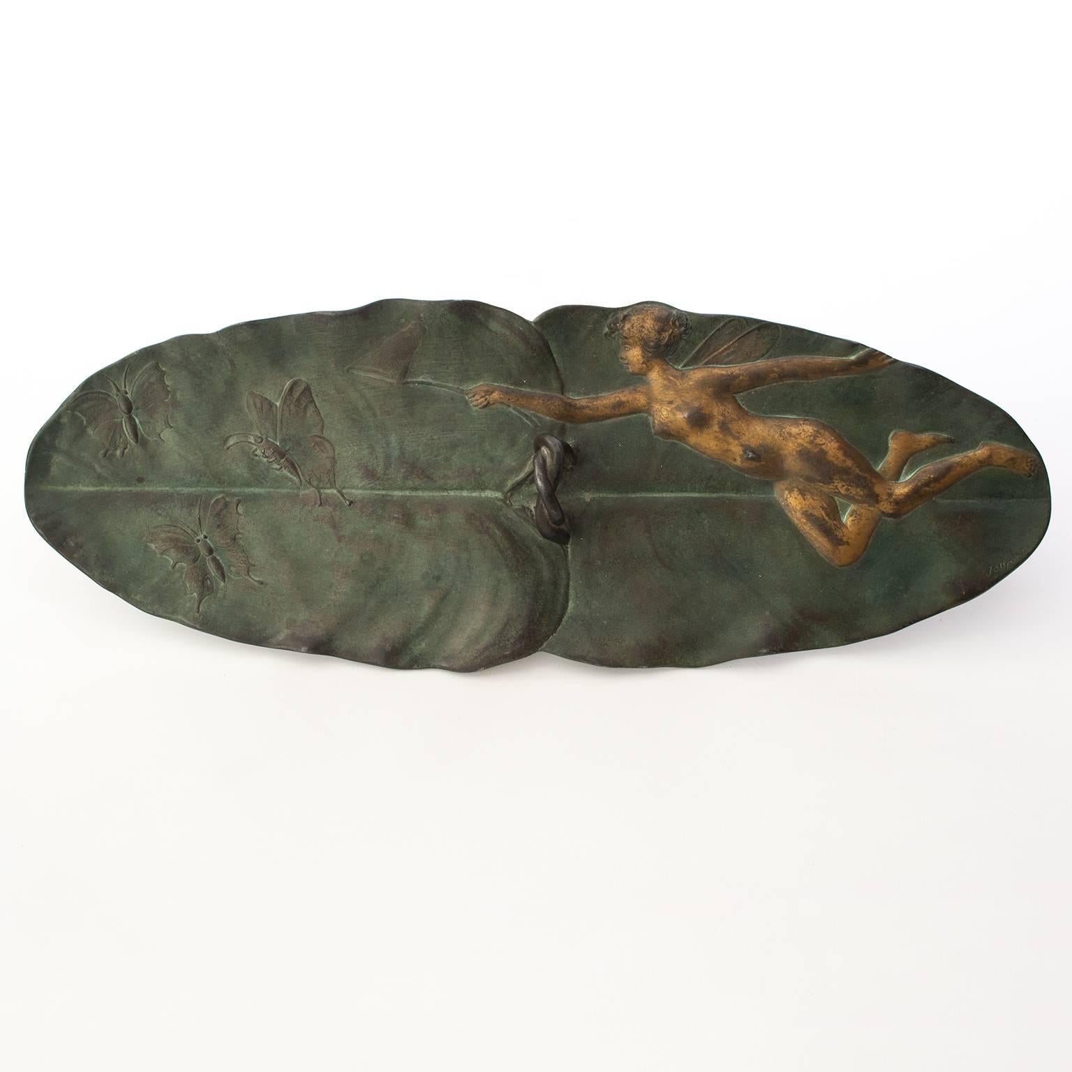 A French Art Nouveau period, painted metal tray in the form of overlapping leaves or lily pads with a center handle. The tray is decorated with a winged female figure in relief holding a wand and is in pursuit of a group of butterflies. Signed