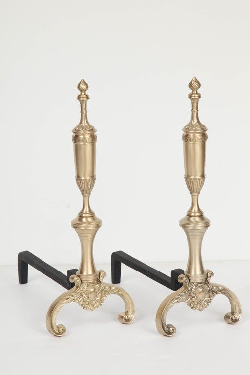 Fantastic set of satin brass andirons with neoclassical style elements which includes a subtle Greek key border, curving body and spiral finials.