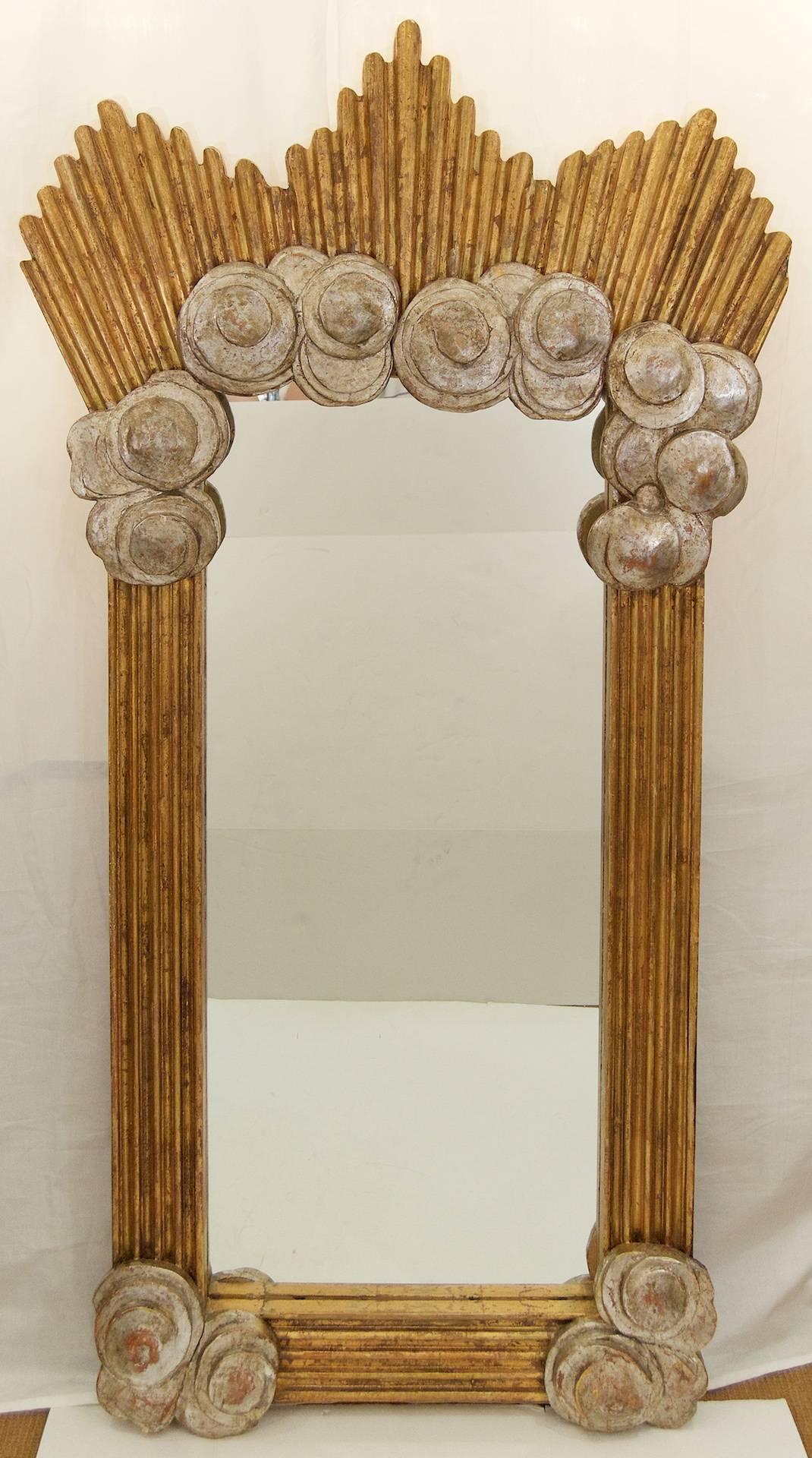 Dramatic and well-sized mirror of American Deco style, the heavily carved wood frame in silver and gold leafed finish. The fluted frame capped by sunburst flares, with large roundel appliques at the corners.

Uncertain age, likely early 3rd