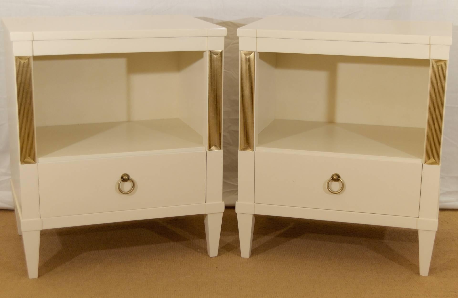 Classically formed nightstands or end tables by John Stuart. Newly relacquered in a satin muslin tone, with gilt and patinaed accents to the pilasters framing the central open storage area. Lower drawer with original brass pulls, patinaed to match
