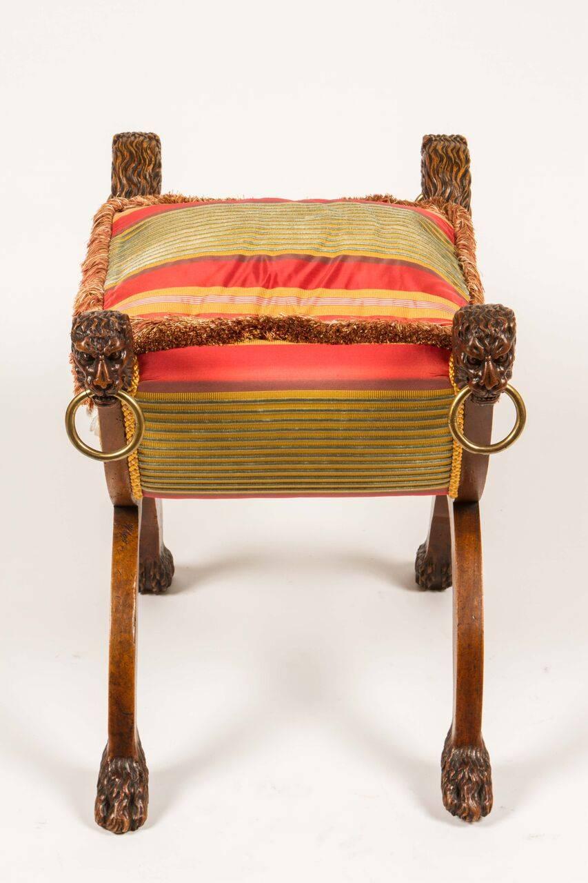 Regency era bench with carved lion's heads with brass rings in their mouths and carved paw feet. Newly upholstered in a Manuel Canovas striped silk and trimmed in a luxurious caterpillar trim with rock crystal florets on each side. This would be