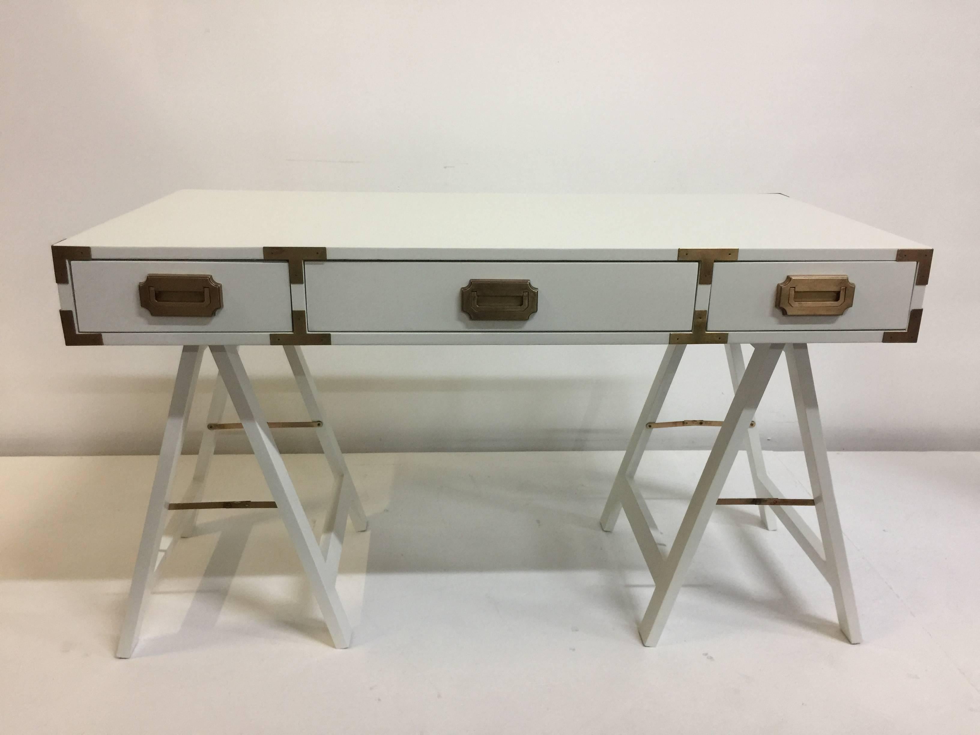 Lacquered in pristine white matte finish, this Campaign style desk sits upon trestle style sawhorse bases with original brass hardware. Three ample drawers open and close perfectly.