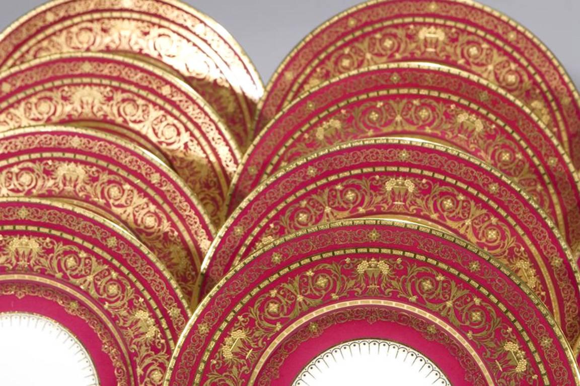 A set of 12 Mintons service, presentation plates with rich burgundy borders are decorated with three bands of acid-etched gold and a neoclassical motif of urns overflowing with grapes. A rare color, these service plates exemplify the fabulous