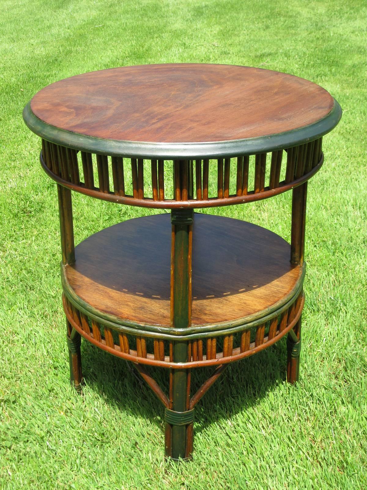 Round stick wicker end table in natural stained finish and faded green painted trim. Wood top and bottom shelf, both having paired reed aprons. Handy portable tea table height. Last photo shown with matching armchairs, listed and priced separately.