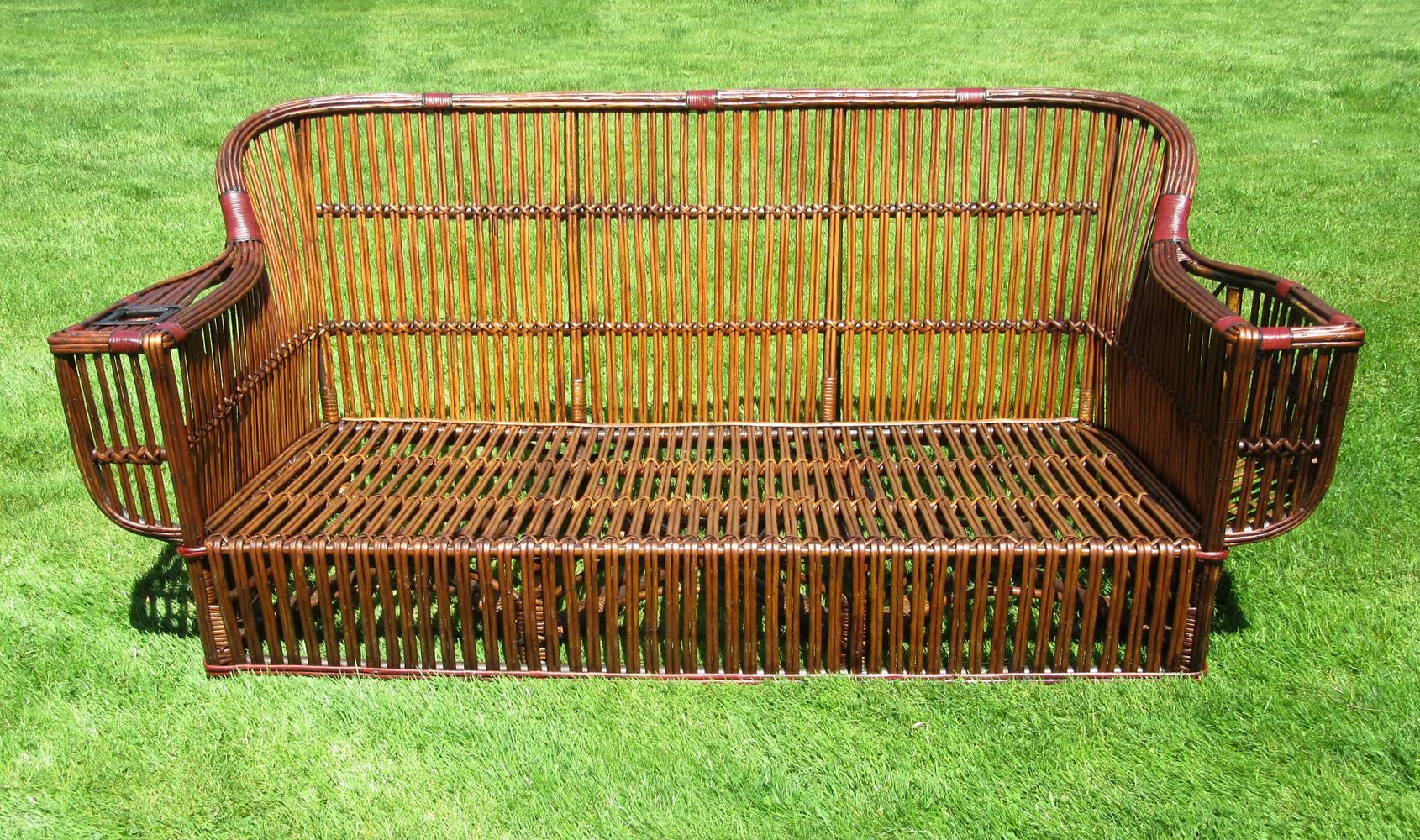 Large-scale stick wicker sofa in medium-toned natural stained finish with red and black painted trim. Deep seated with closely paired rattan strands overall. Large framed armrests having magazine rack and beverage holder.