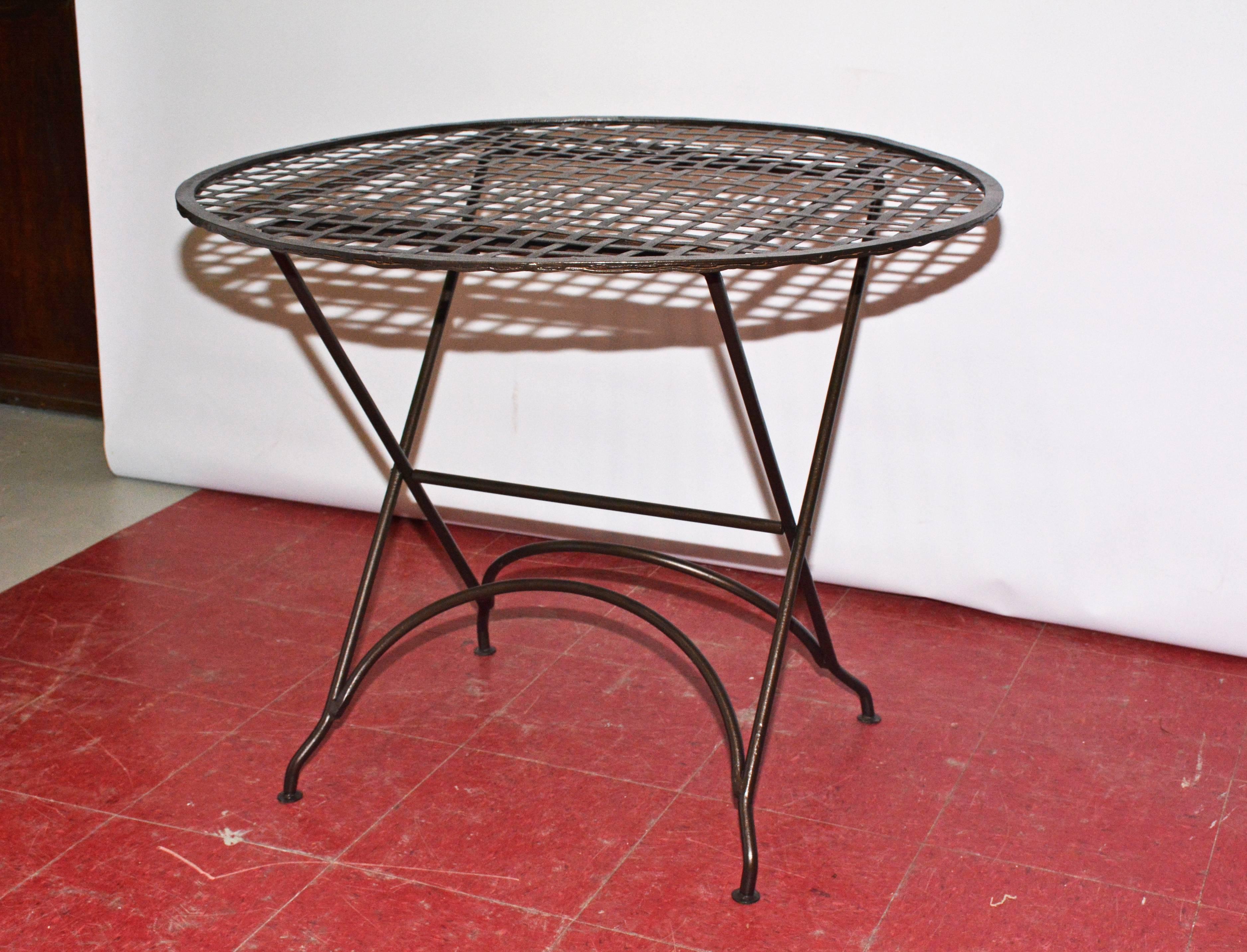 Indoor or outdoor bistro table with cross-hatch banded tabletop of iron attached to a rim. The top of one side of the criss-cross legs can be unhooked from underneath the tabletop and folded flat while the top can be laid flat against the legs. Can