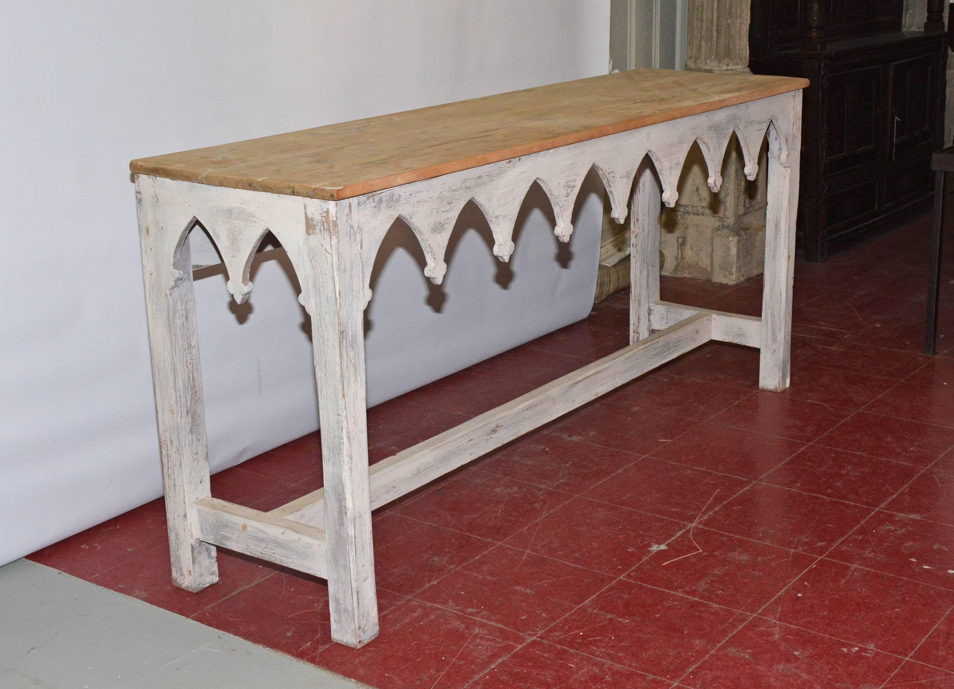 Handcrafted in the Regency-style, the sturdy rustic console or sofa table has an apron of neo-gothic arches and legs painted white and a sanded wood top.