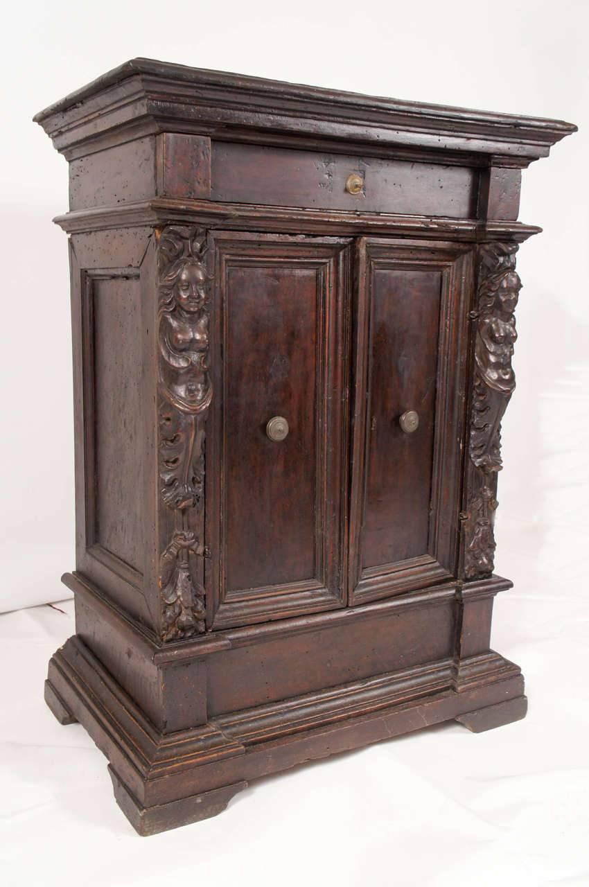Small in size but impressive in the scale of its architectural moldings and details, this Italian Renaissance-style cabinet contains two shelves behind double paneled doors and a center drawer above. Hand-carved caryatids and acanthus leaves flank