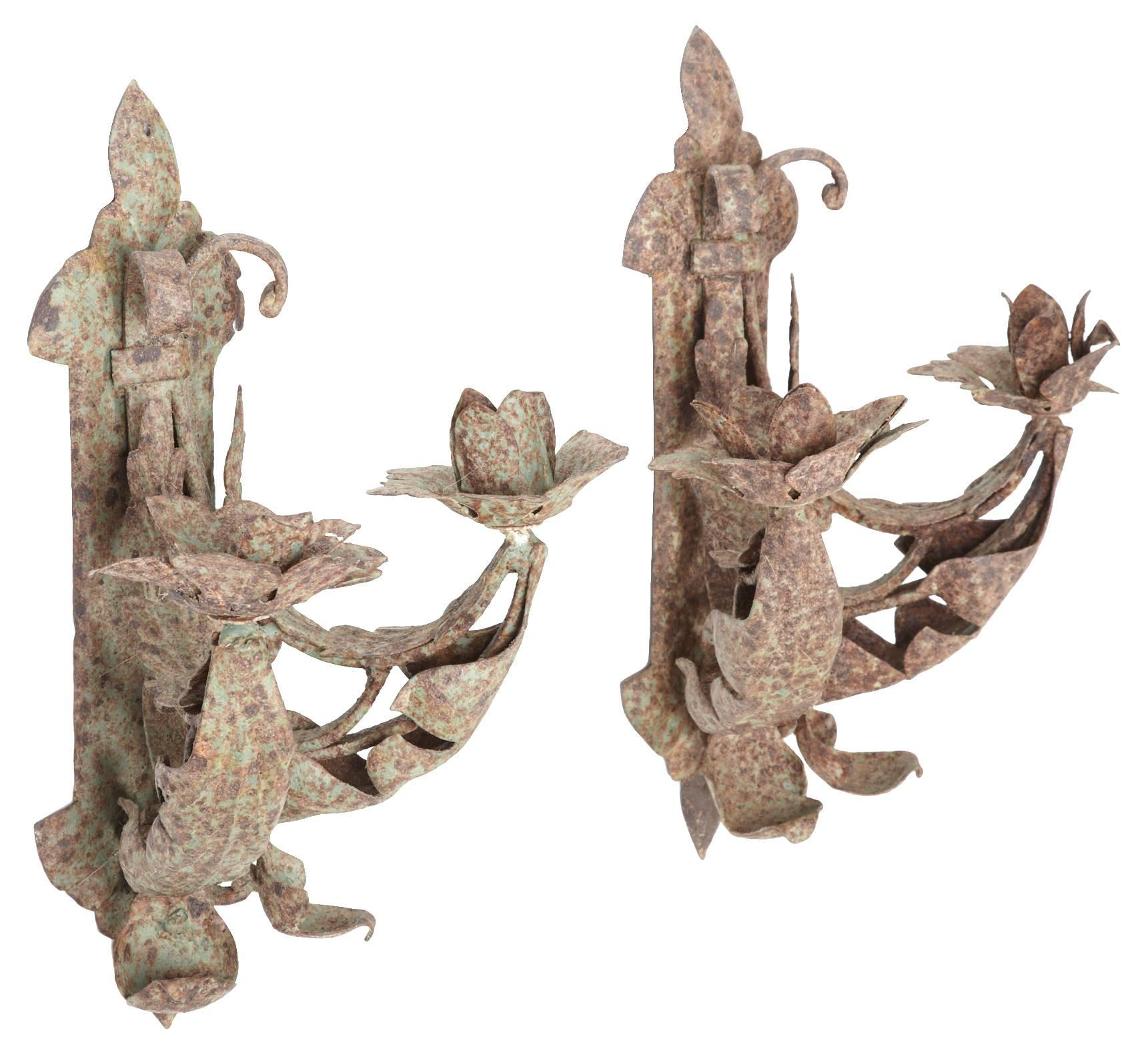 The pair of antique handcrafted iron sconces are decorated with various forms of leaves and are secured by brackets, each with a hole at the top for attaching a screw or nail to a wall. Each sconce holds two candles.