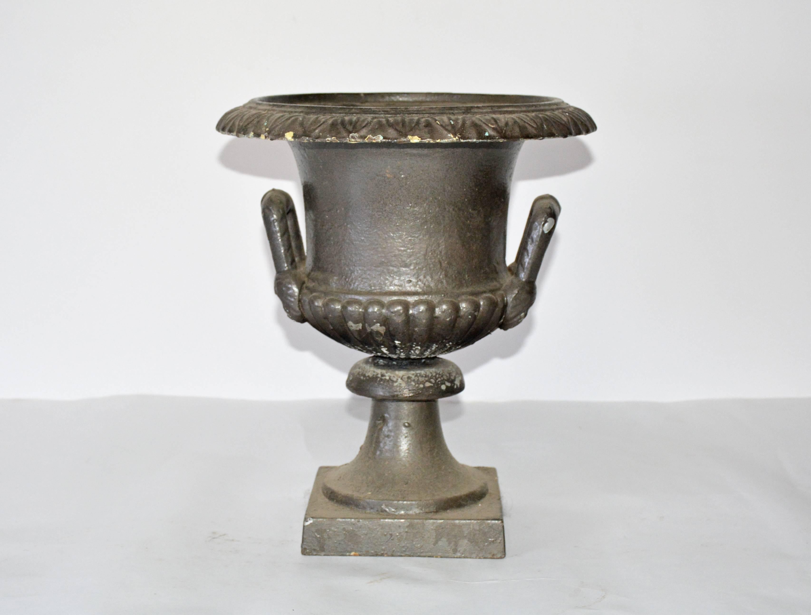 The antique classical garden urn is made of cast iron and has two handles and a flared rim. The container has a fluted design encircling its bottom. The handles are screwed to the container, while the latter is screwed to the bottom.