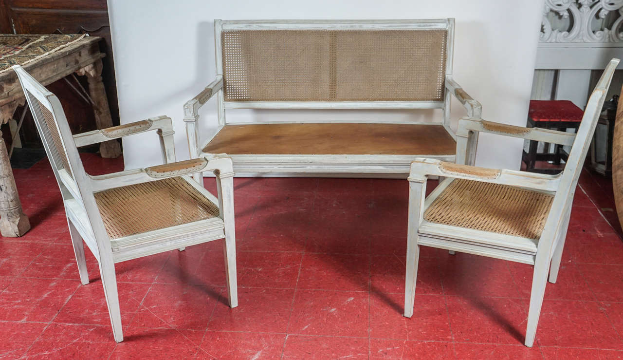 This Directoire or Gustavian style trio are perfect for a hall, enclosed loggia or sun porch. The armchairs have cane backs and seats. The bench has a cane back and wood seat. All are painted rubbed French grey and require seat cushions and arm pads