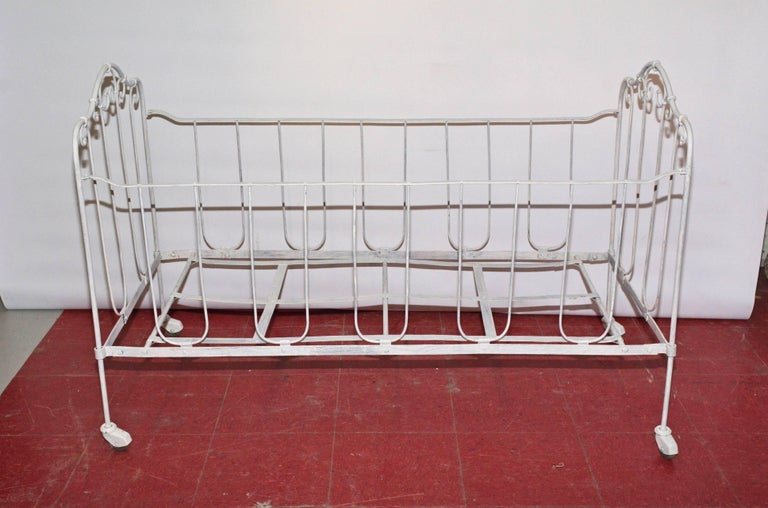 This hand worked wrought iron crib rolls on casters for easy movability. The slatted bottom will hold a mattress and bumpers may be tied to the vertical supports. The end framing is decorated with curlicues. The crib is newly painted white. Front