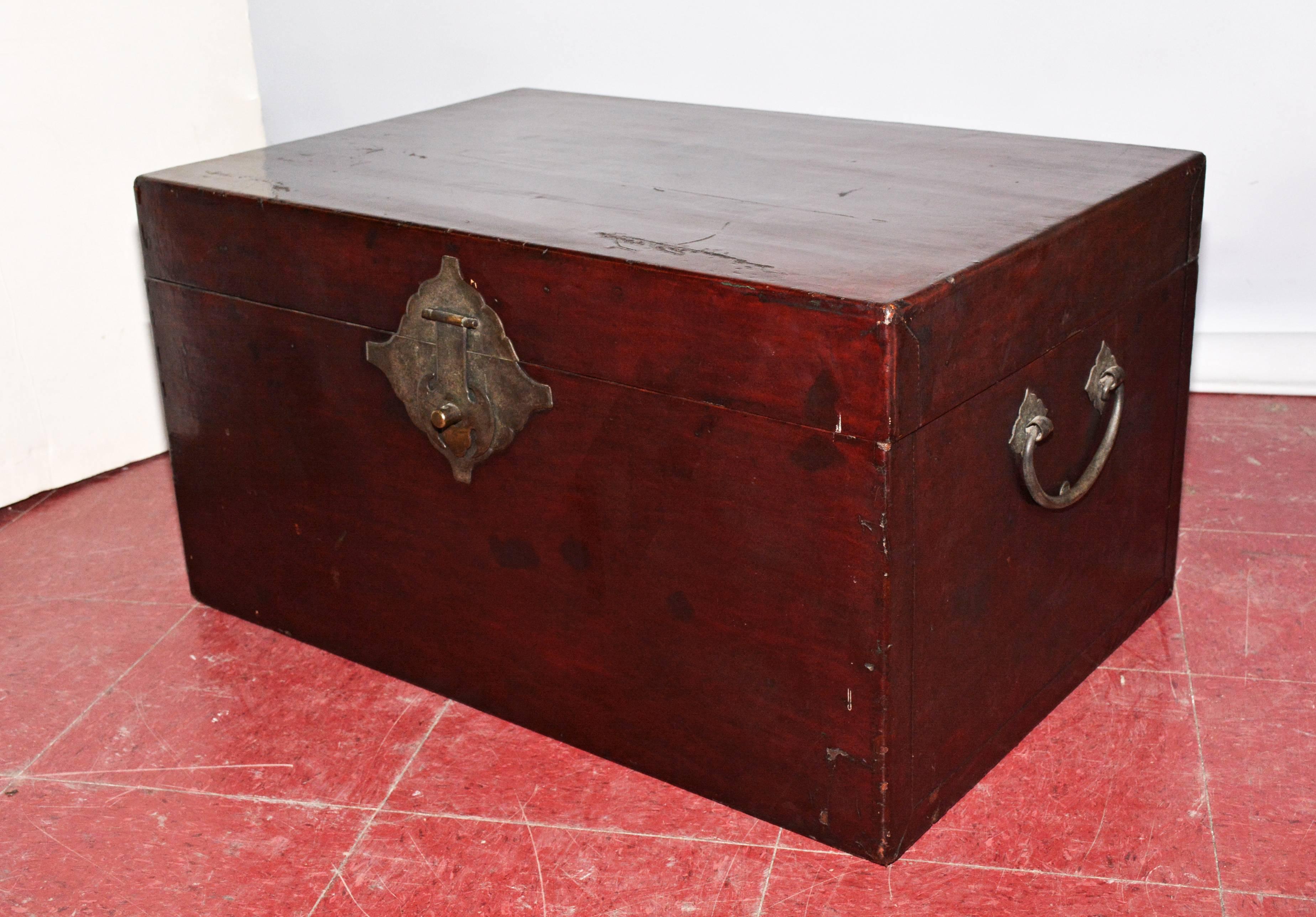 The Chinese chest or blanket chest has dark reddish brown lacquered wood inside and out, a brass latch, and a pair of iron handles. The sides are edged with thin sheets of overlapping wood having imitation stitching. The lid is attached to the