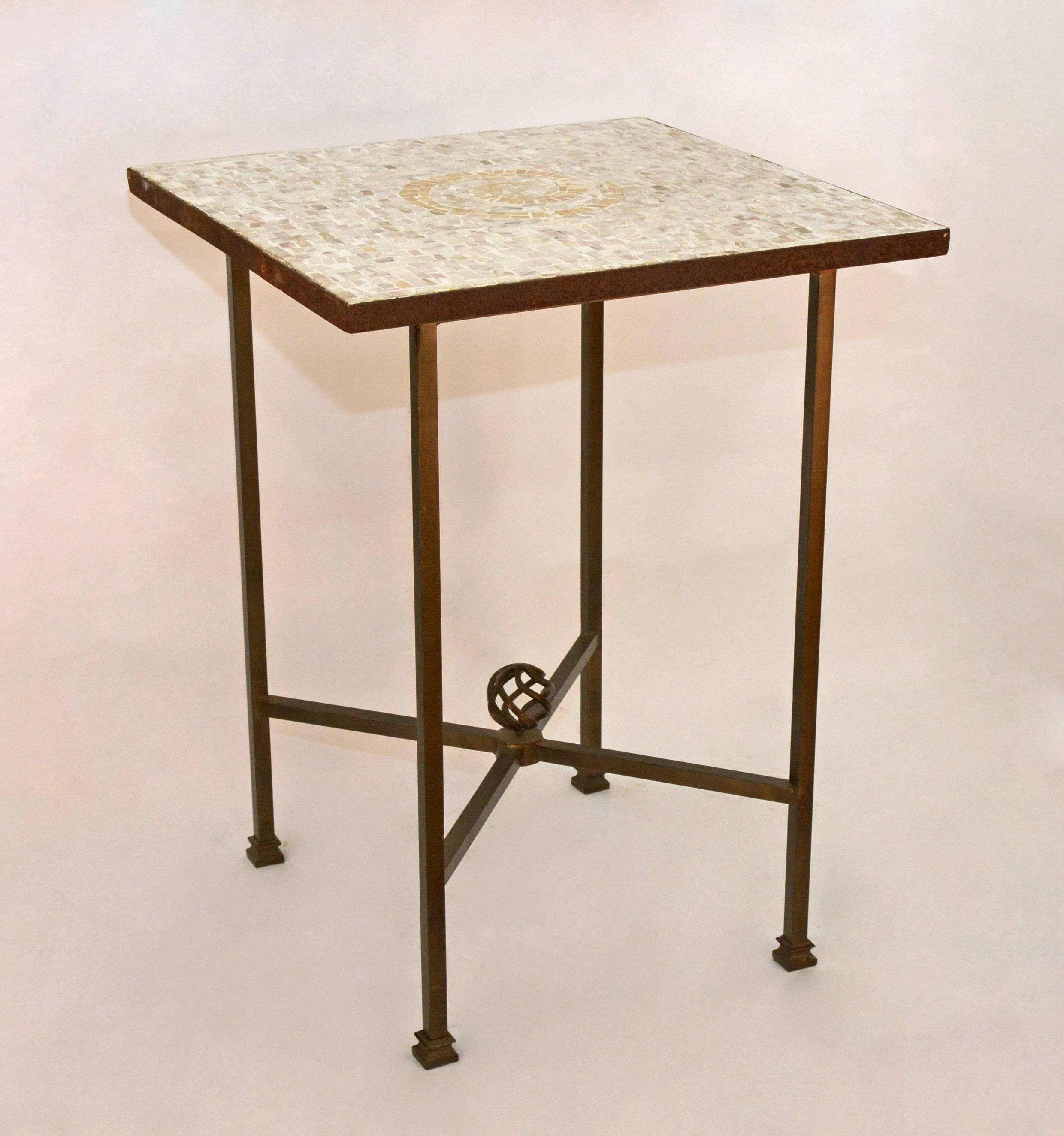 The mosaic top has inset tiles in pale shades of white, gray beige and taupe set within a iron square frame. The center design is a spiral shape in tan. The square bronzed colored base has criss-cross stretchers with a spiral finial in the center