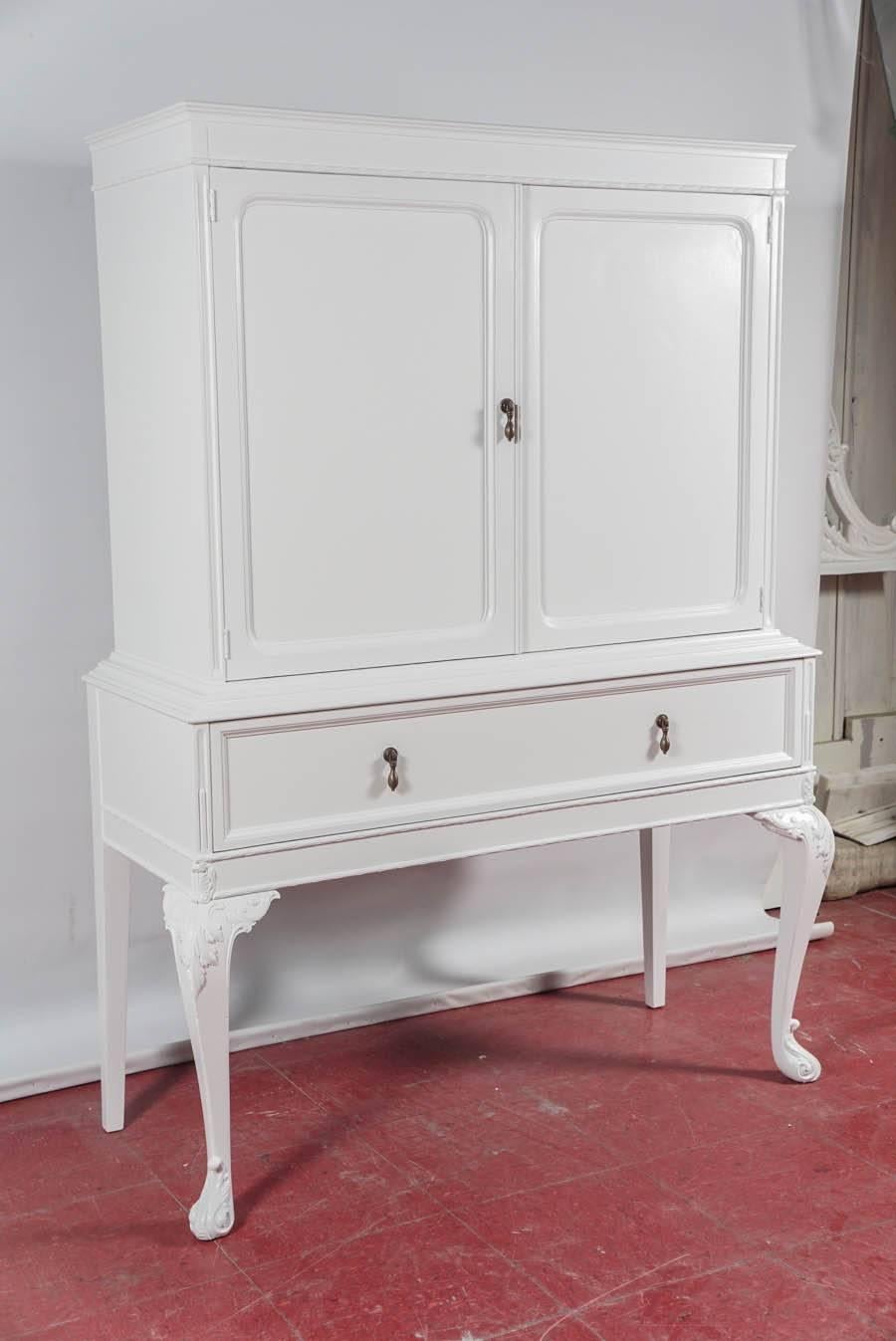 Freshly painted white, the early 20th century china cabinet has one generous size dovetail drawer and two shelves behind a pair of paneled doors. One shelf has slots for angling plates.